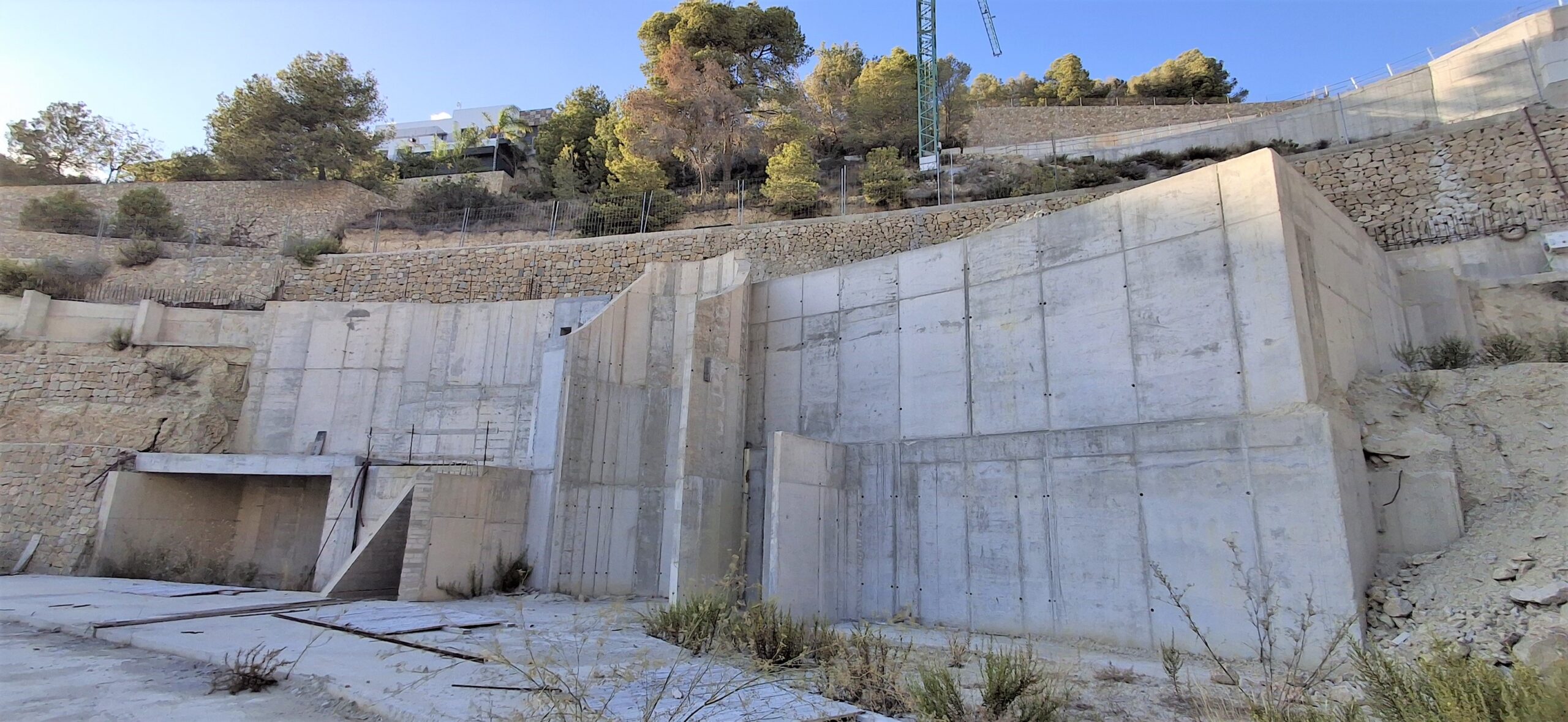 Vue des murs de soutènement en béton armé et des terrasses en pierre intégrés dans un terrain en pente avec une végétation méditerranéenne.