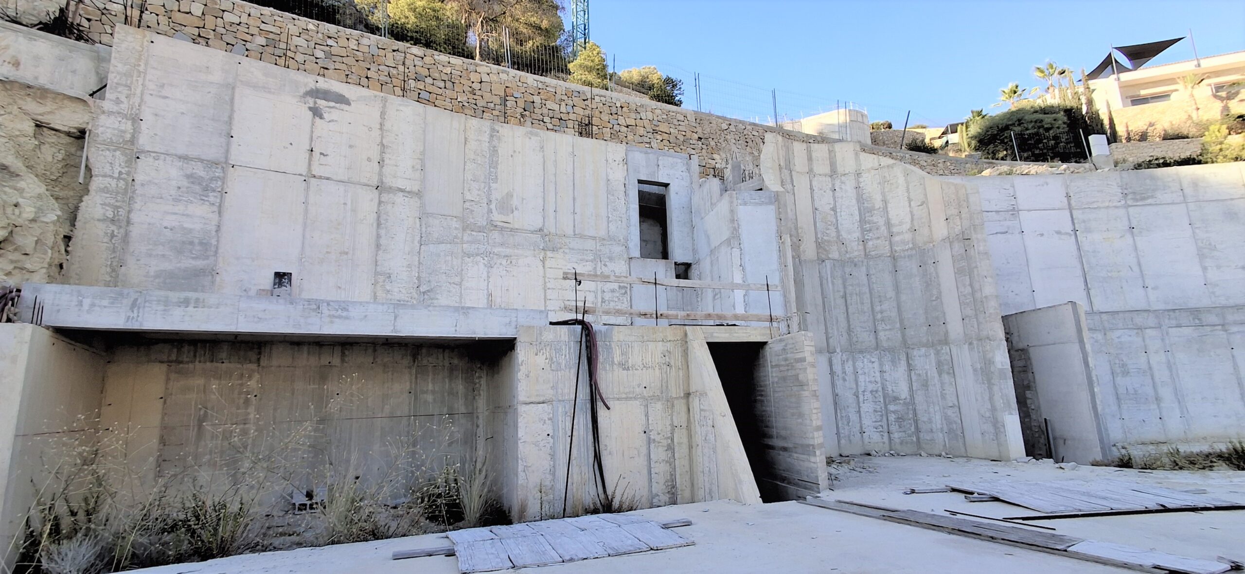 Vue structurelle des murs de soutènement en béton armé, avec maçonnerie en pierre intégrée au niveau supérieur et ouvertures architecturales sur un terrain en pente.