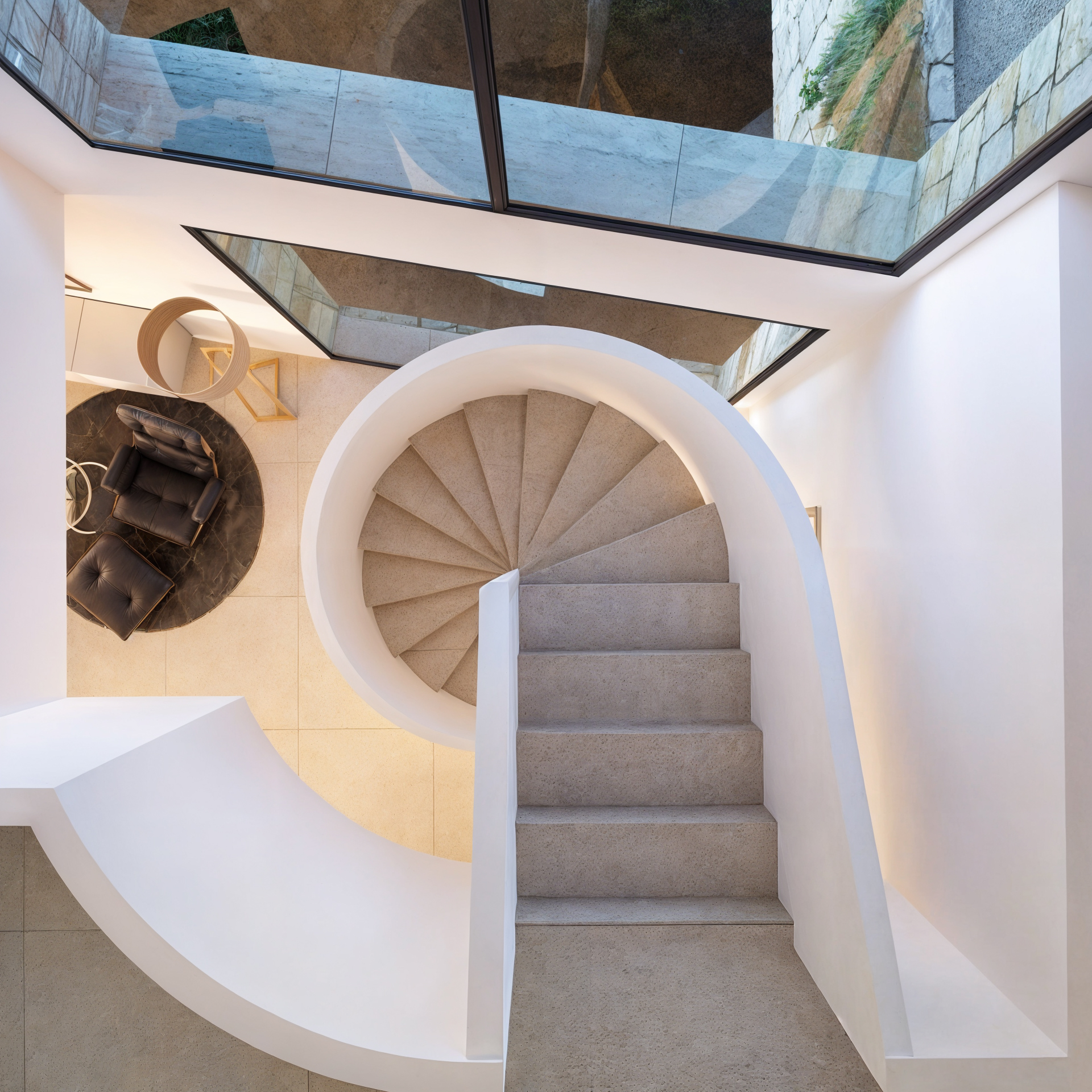 Escalier en colimaçon en pierre avec balustrades blanches courbes, vue aérienne montrant de grandes baies vitrées et un salon avec fauteuil en cuir.
