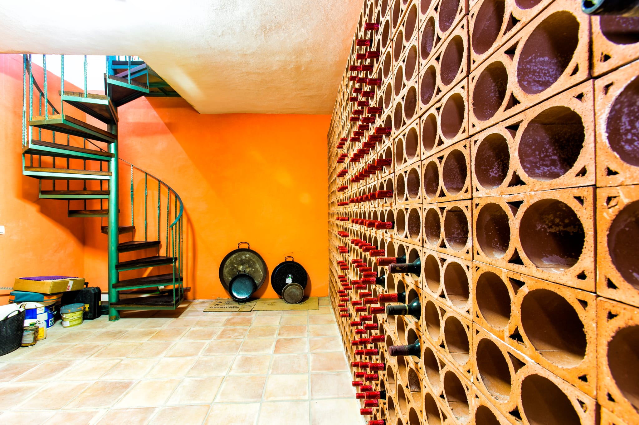 Cave à vin avec casier en terre cuite, mur d'accent orange et escalier en colimaçon en métal vert doté de marches en bois sombre.