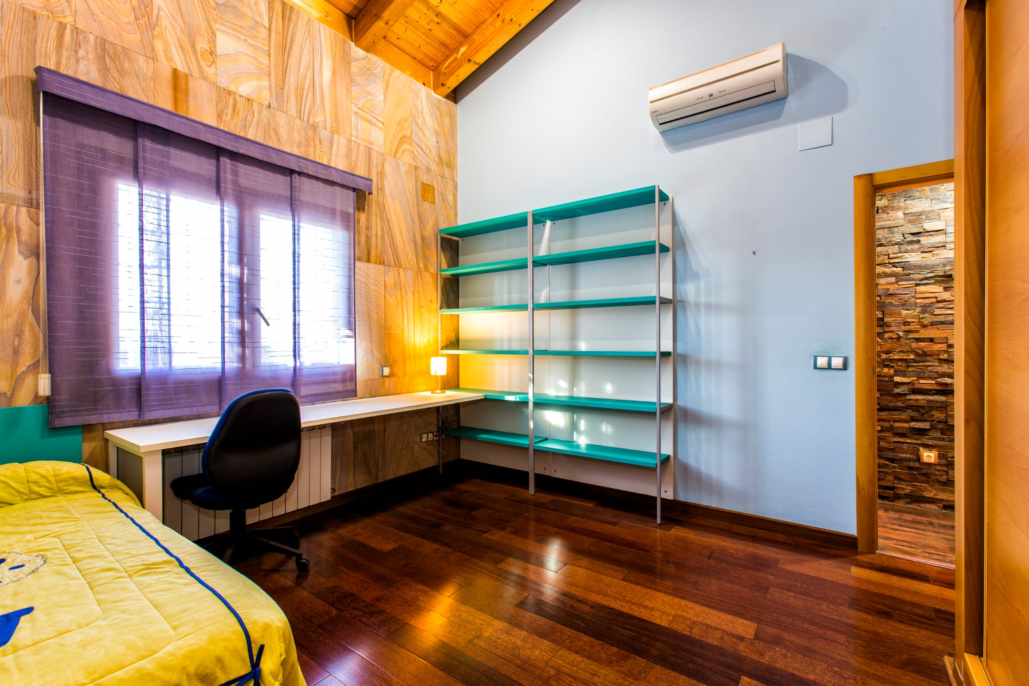 Chambre avec coin bureau, plafonds en bois mansardés, étagère turquoise et bureau blanc sous une fenêtre avec stores violets.