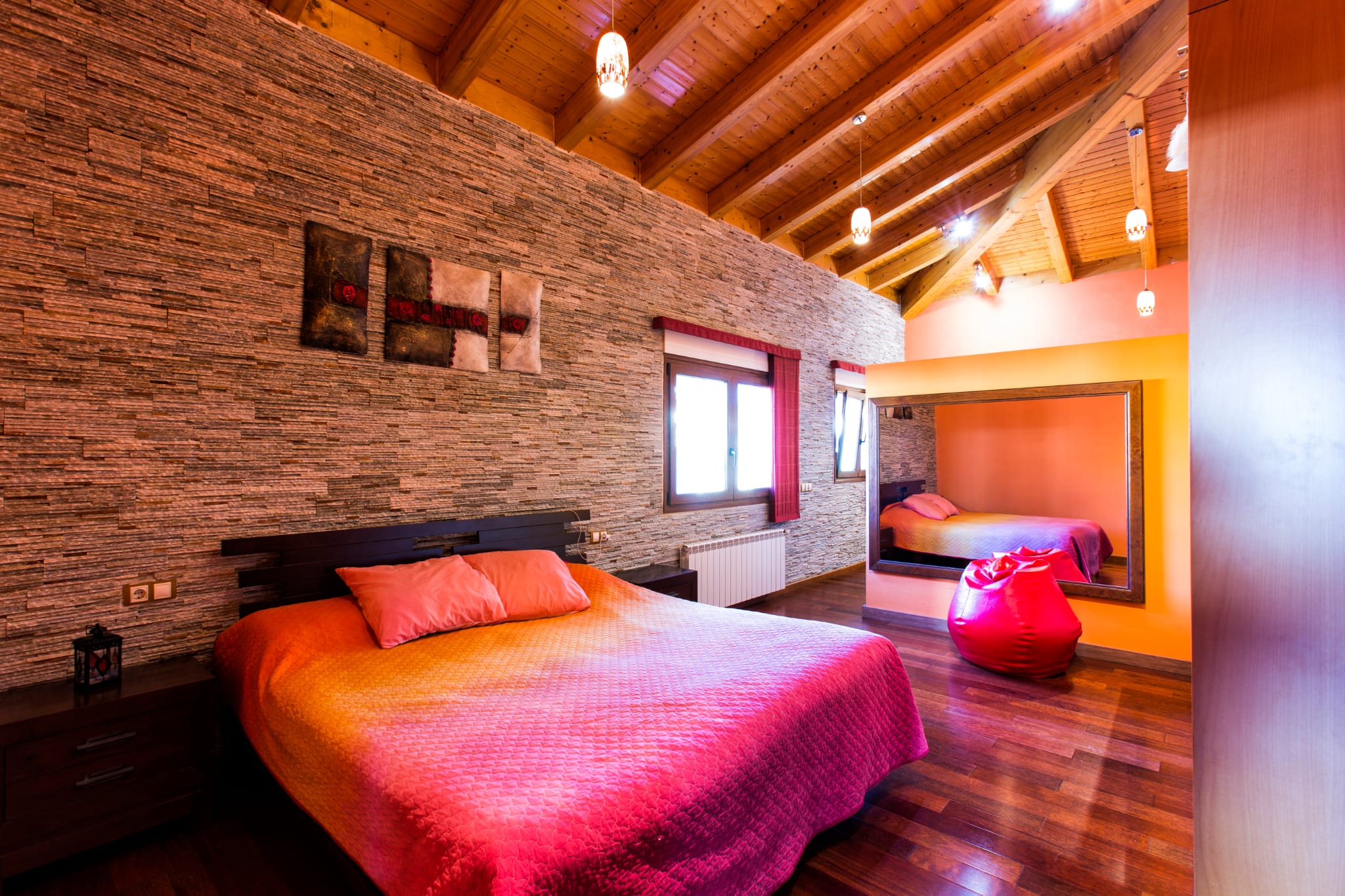Chambre avec mur en pierre texturée, plafond à poutres apparentes en bois et parquet sombre. Comprend un grand miroir mural et des suspensions lumineuses.