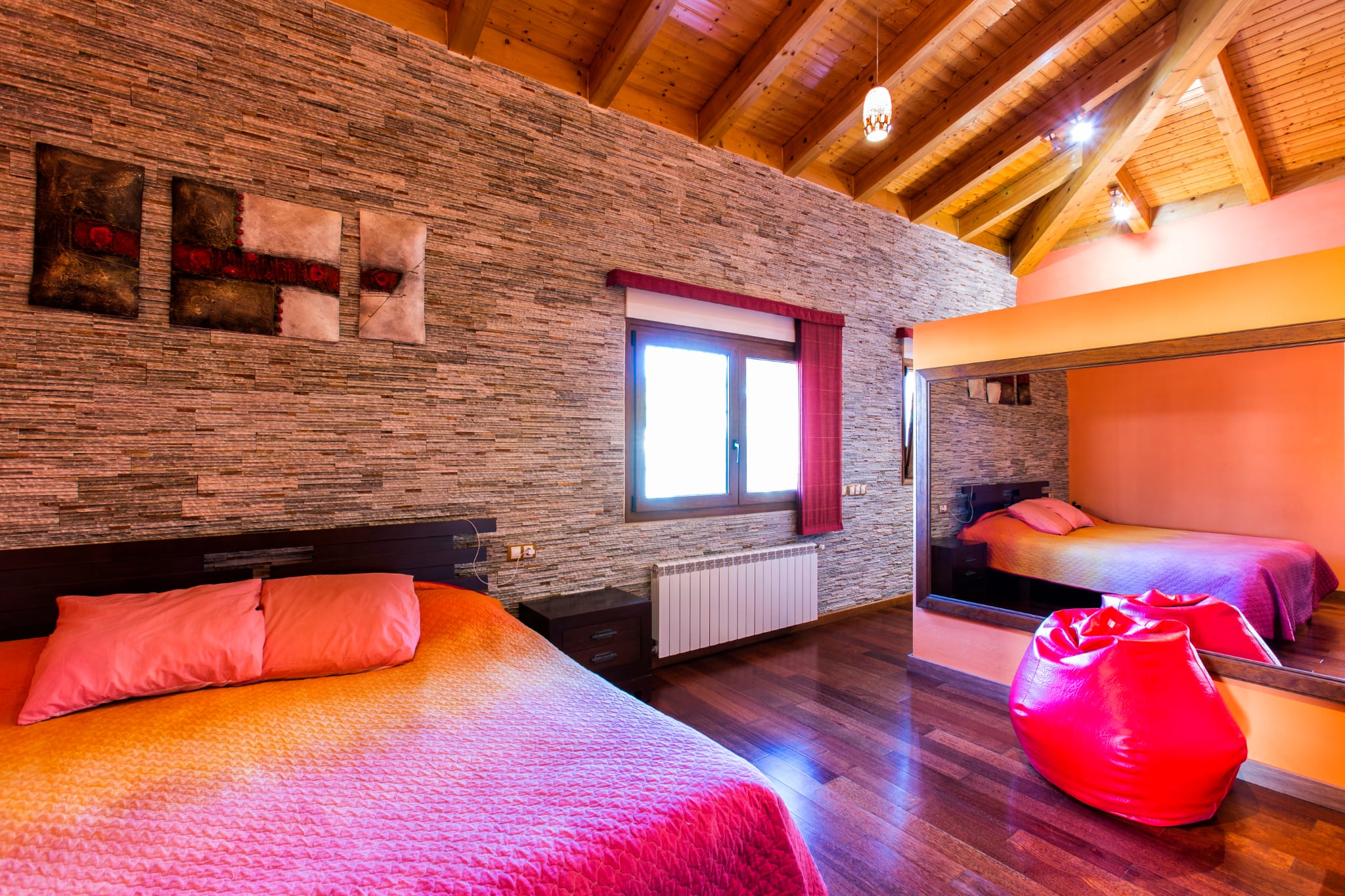 Chambre avec mur en pierre texturée, plafond voûté en poutres de bois et parquet sombre. Un grand miroir mural reflète le lit aux teintes roses et orangées.