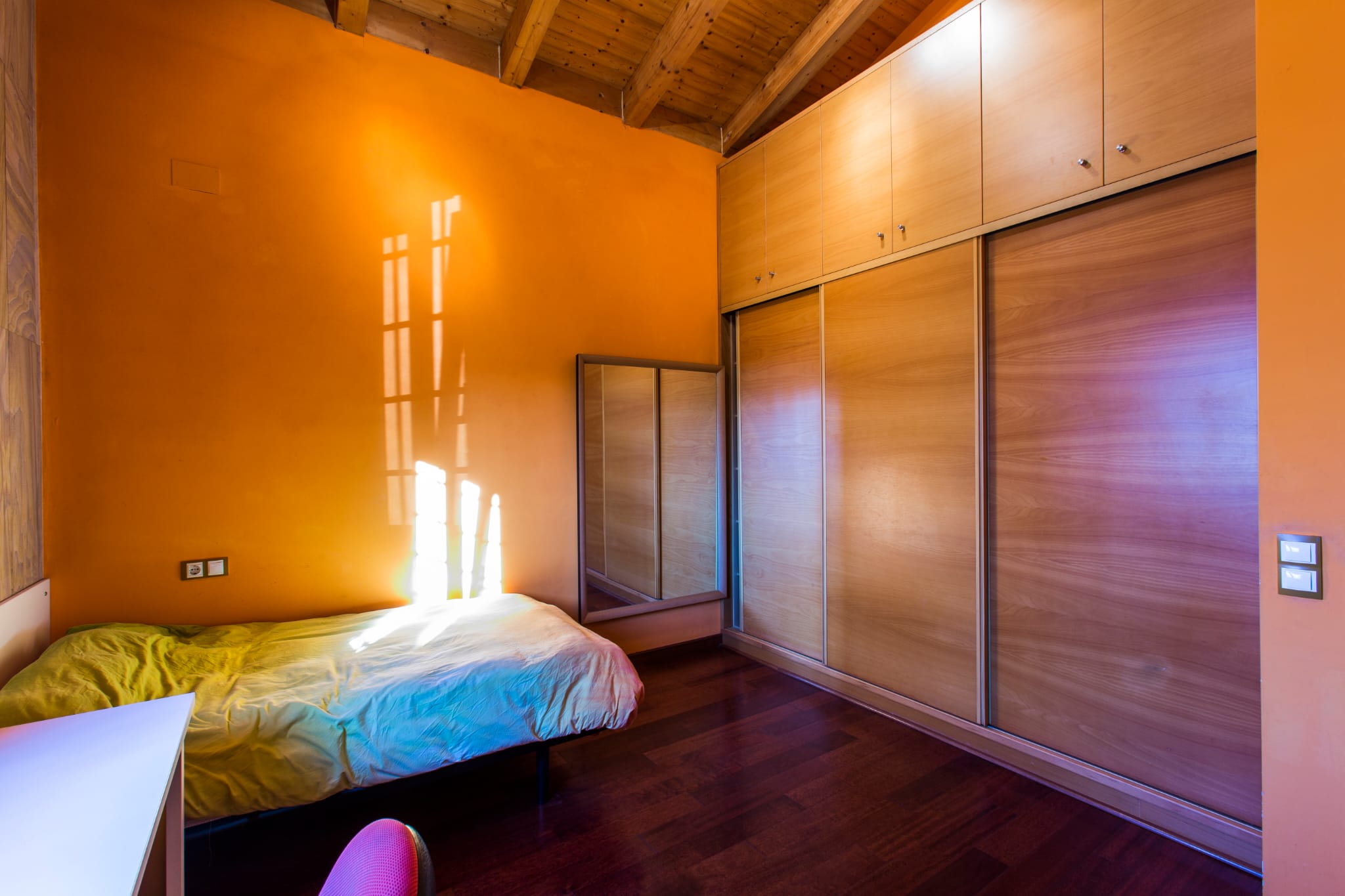 Chambre avec murs orange, plafond en bois à poutres apparentes et une grande armoire encastrée en bois avec portes coulissantes.