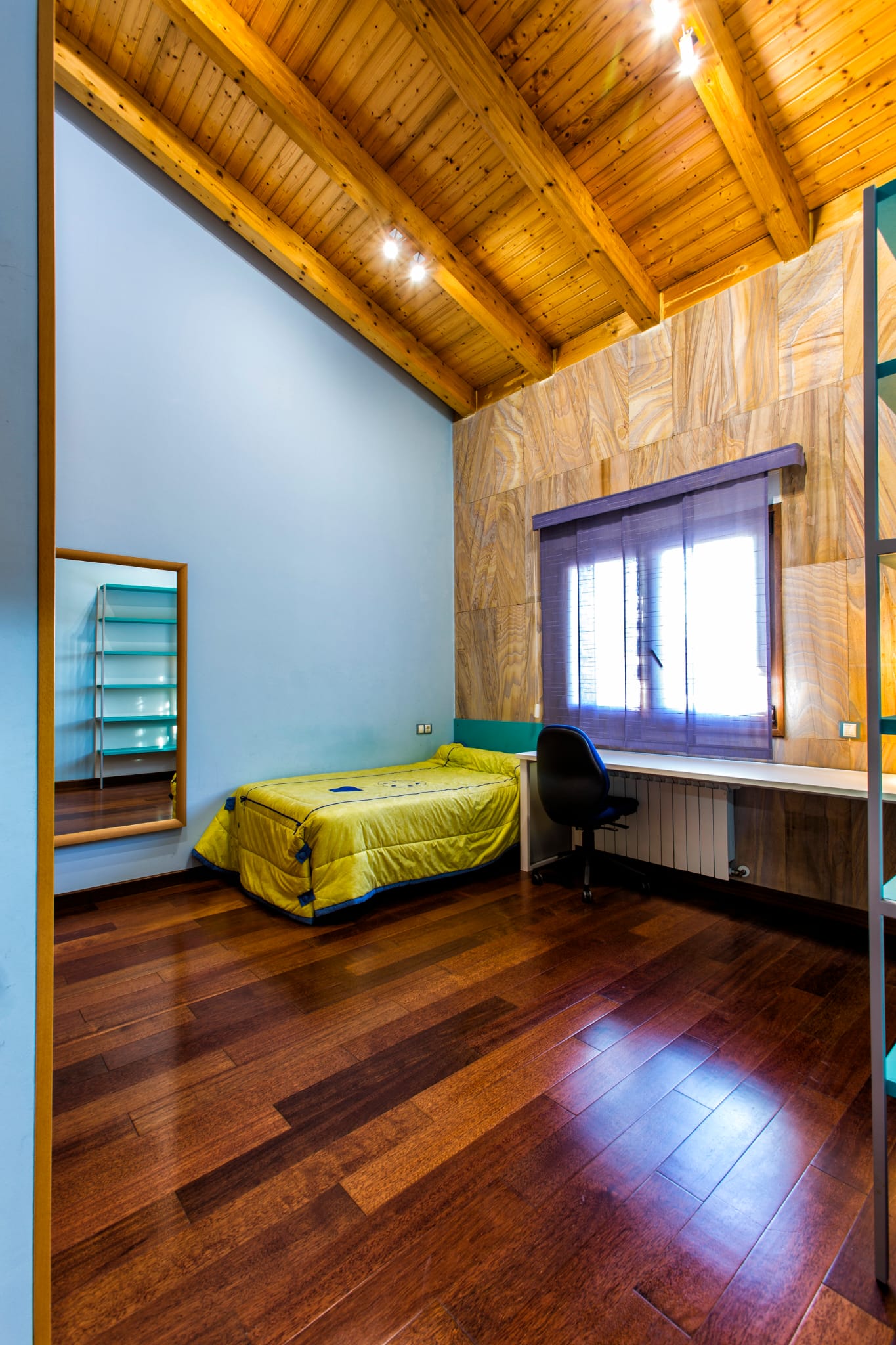 Chambre avec plafond en poutres de bois apparentes, parquet sombre, bureau intégré sous la fenêtre et mur d'accent aux panneaux texturés.