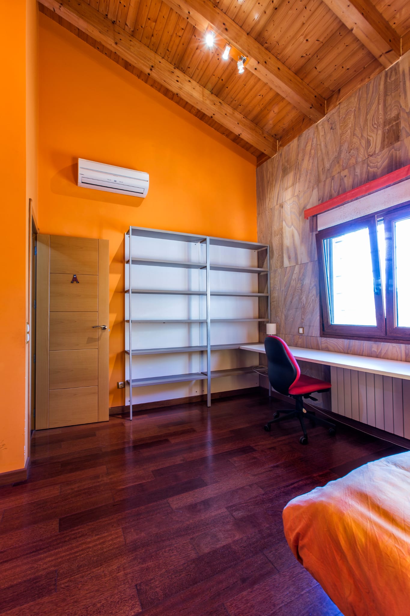 Chambre avec plafond voûté en bois, murs orange, espace bureau intégré avec étagères métalliques, bureau blanc et parquet en bois foncé.