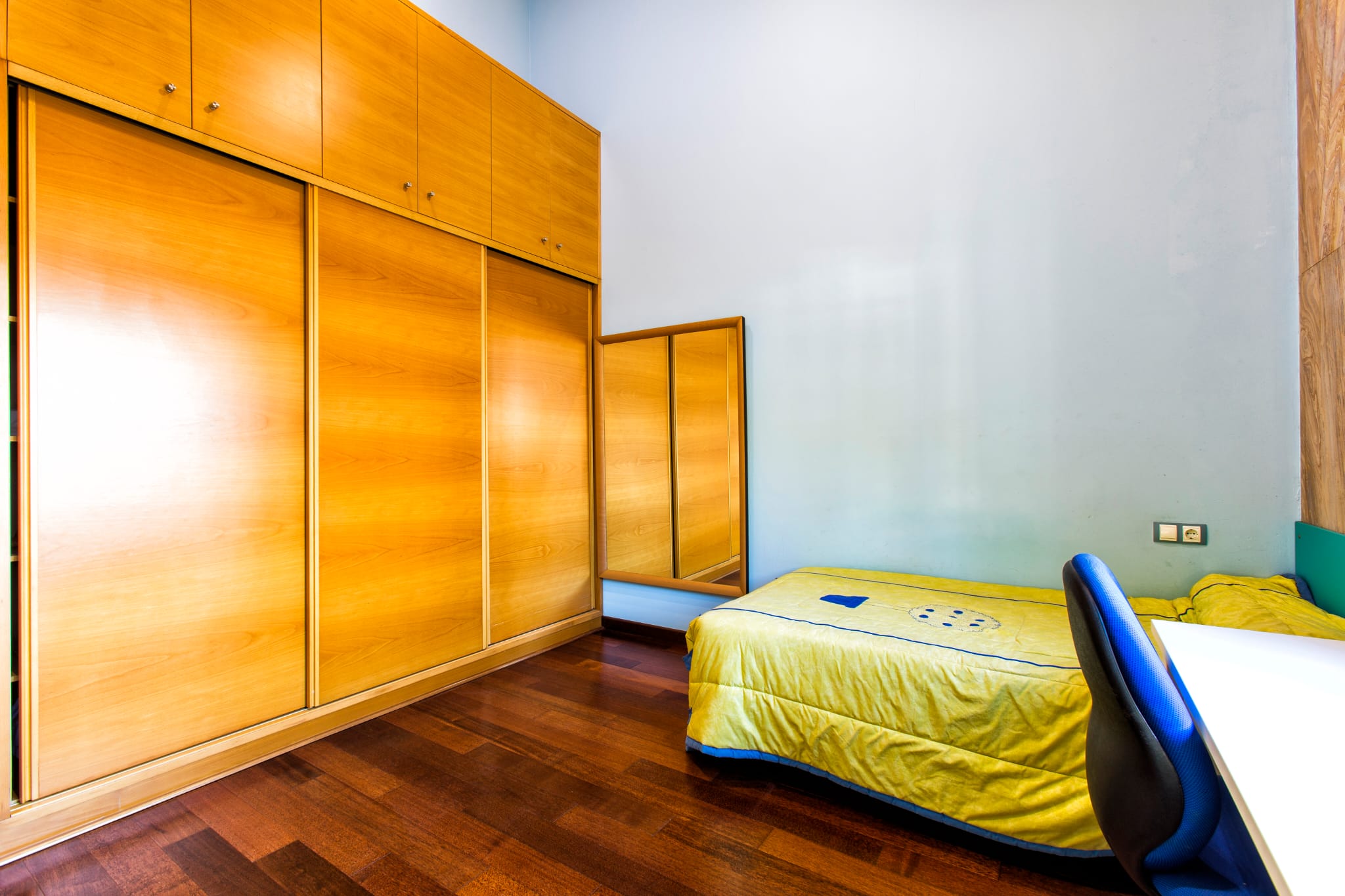 Chambre avec une grande armoire encastrée en bois à portes coulissantes, parquet foncé et lit simple à côté d'un espace de travail.