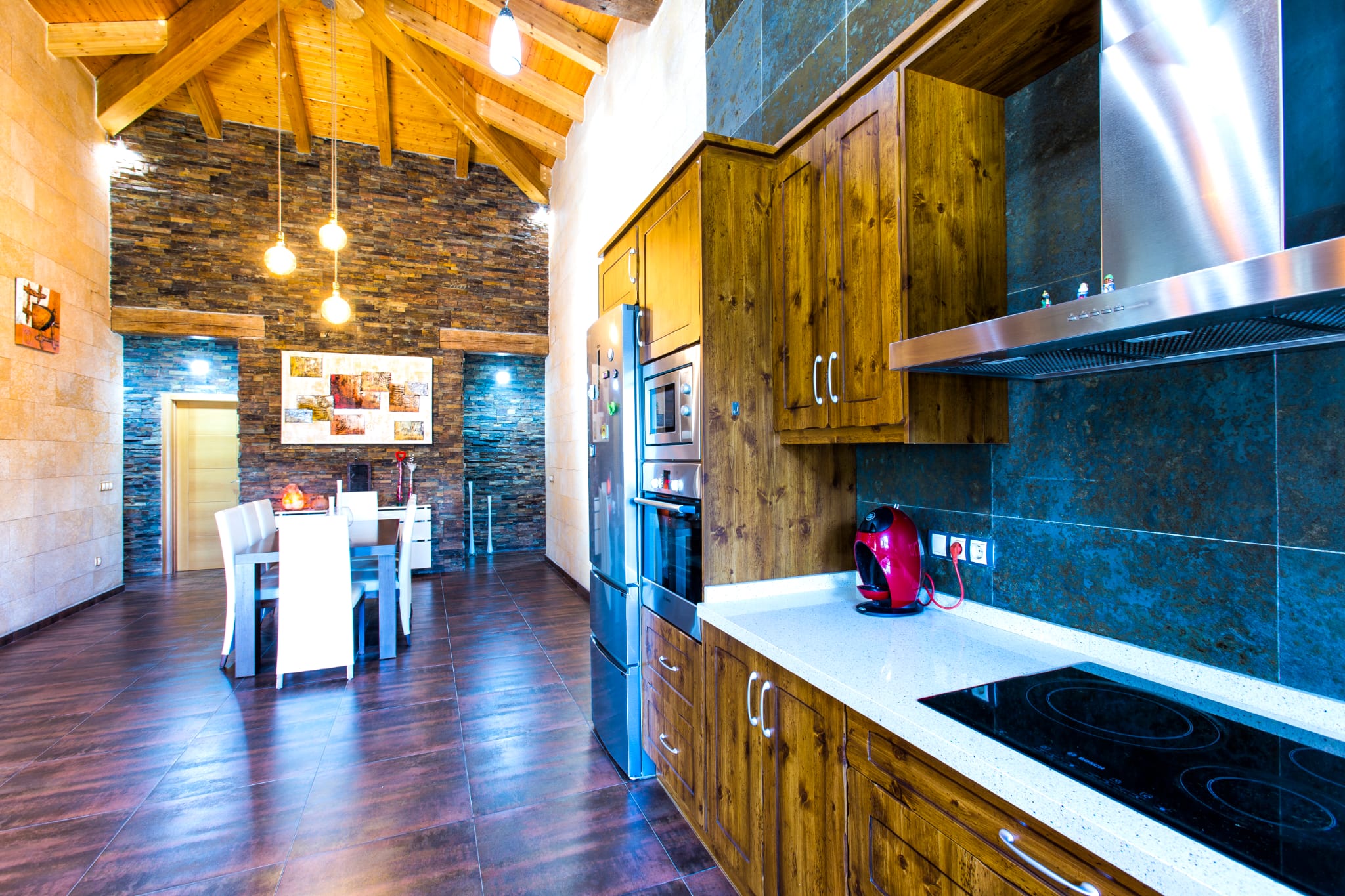 Cuisine équipée de meubles en bois rustique, plan de travail blanc et électroménager intégré, ouverte sur une salle à manger aux murs en pierre et plafond voûté.