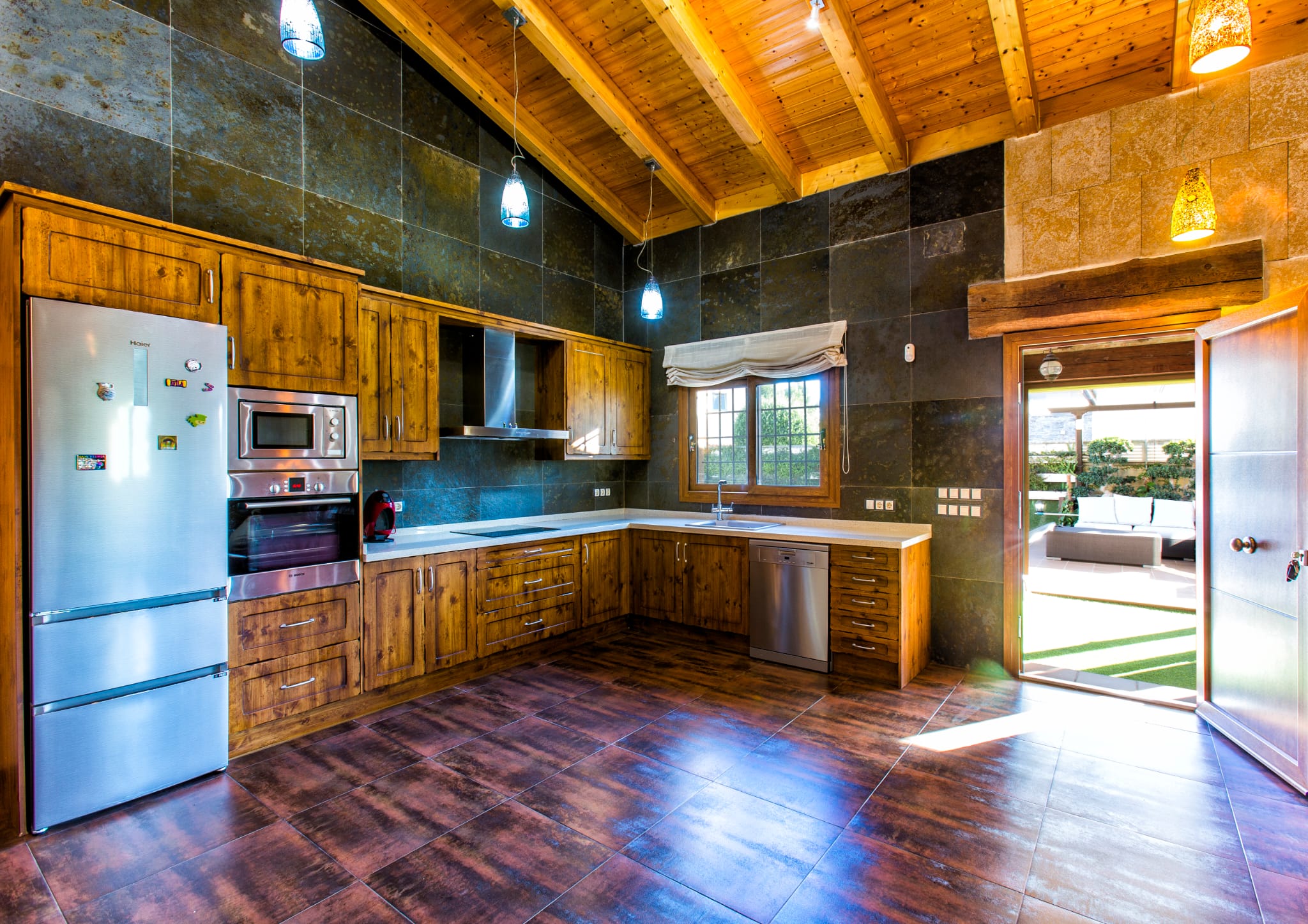 Cuisine rustique avec meubles en bois, murs en ardoise sombre et plafond à poutres apparentes. Équipée d'appareils en inox et ouvrant sur une terrasse extérieure aménagée.