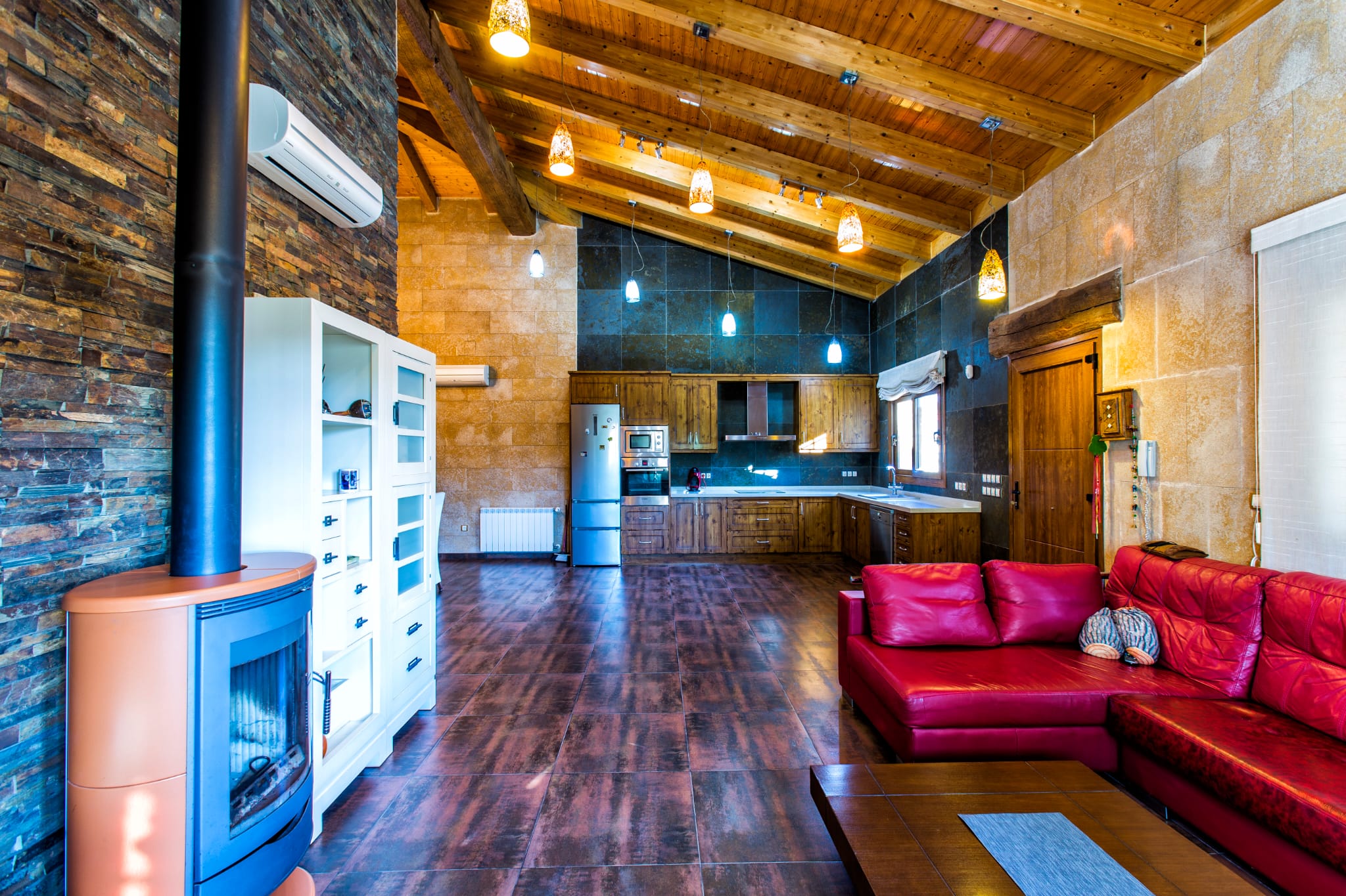 Espace de vie ouvert avec plafond à poutres apparentes, poêle à bois moderne et cuisine rustique équipée, mettant en valeur des murs en pierre et un sol en grès.