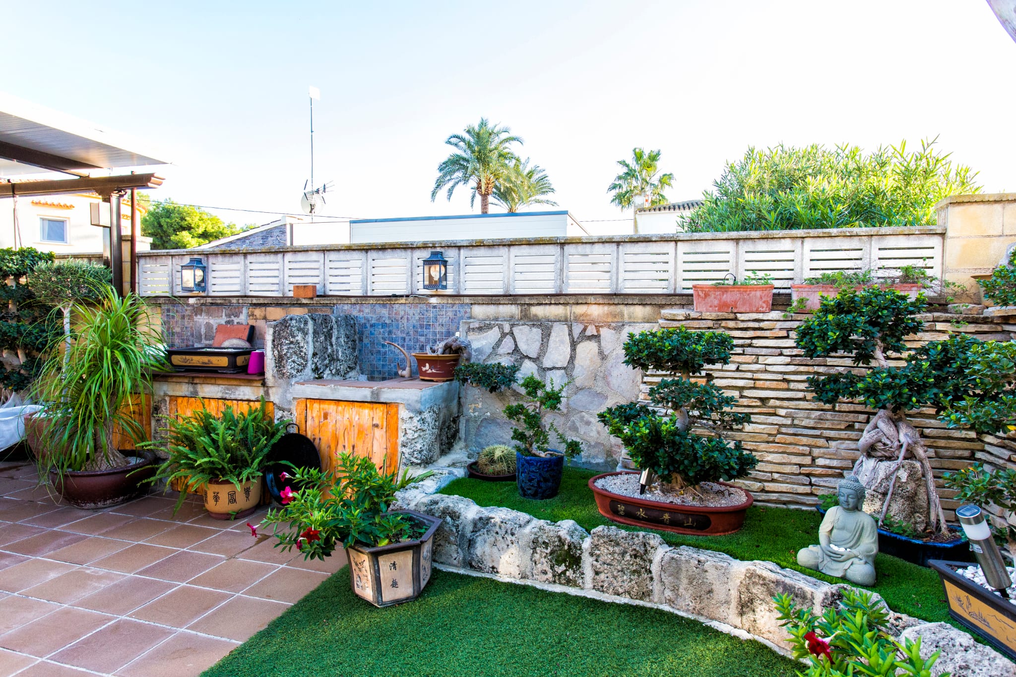 Jardin patio de style zen avec gazon artificiel, murs en pierre sèche et collection de bonsaïs. Comprend un comptoir en pierre avec mosaïque bleue.