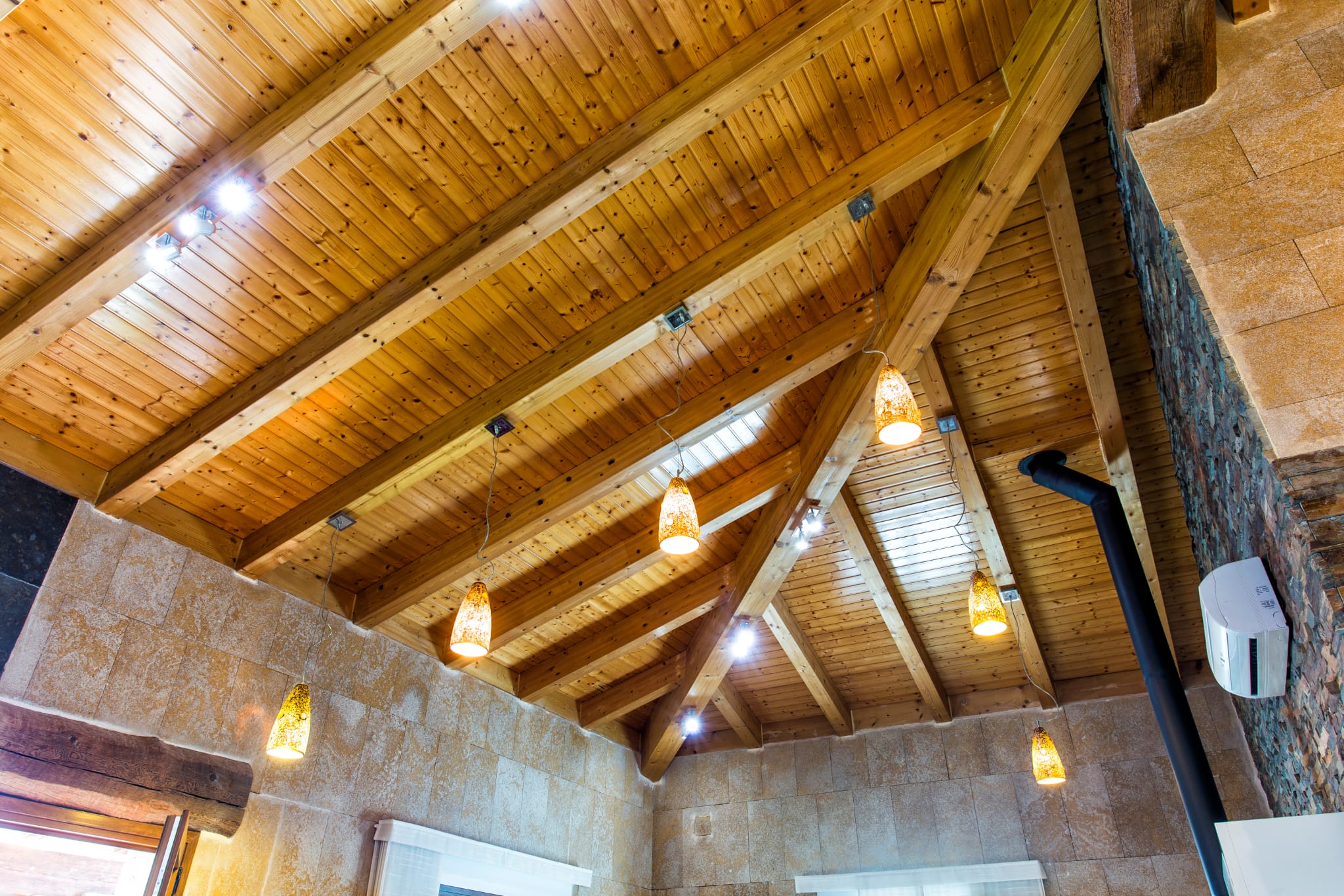 Plafond cathédrale avec poutres apparentes en bois et lambris de pin, agrémenté de suspensions en verre ambré et de murs revêtus de pierre.