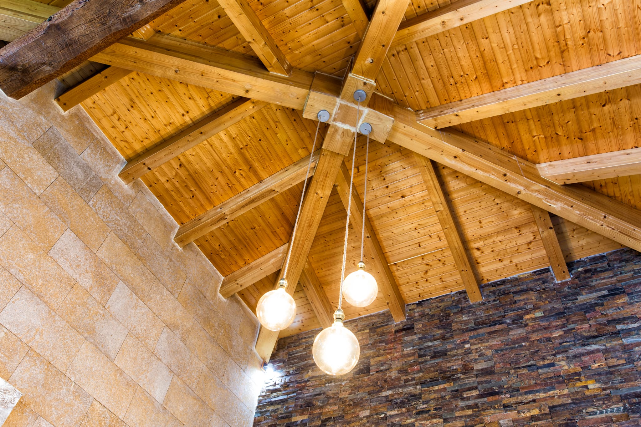 Plafond voûté avec poutres en bois apparentes en étoile, éclairé par trois suspensions sphériques sur des murs en pierre de taille et parement sombre.