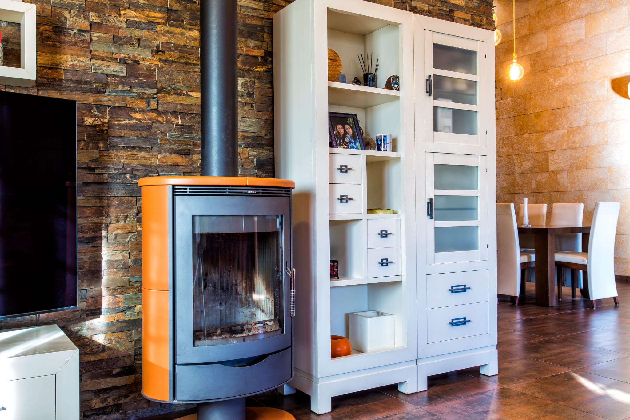 Poêle à bois moderne avec finitions orange sur mur en pierre naturelle, jouxtant un buffet blanc et un espace salle à manger carrelé.