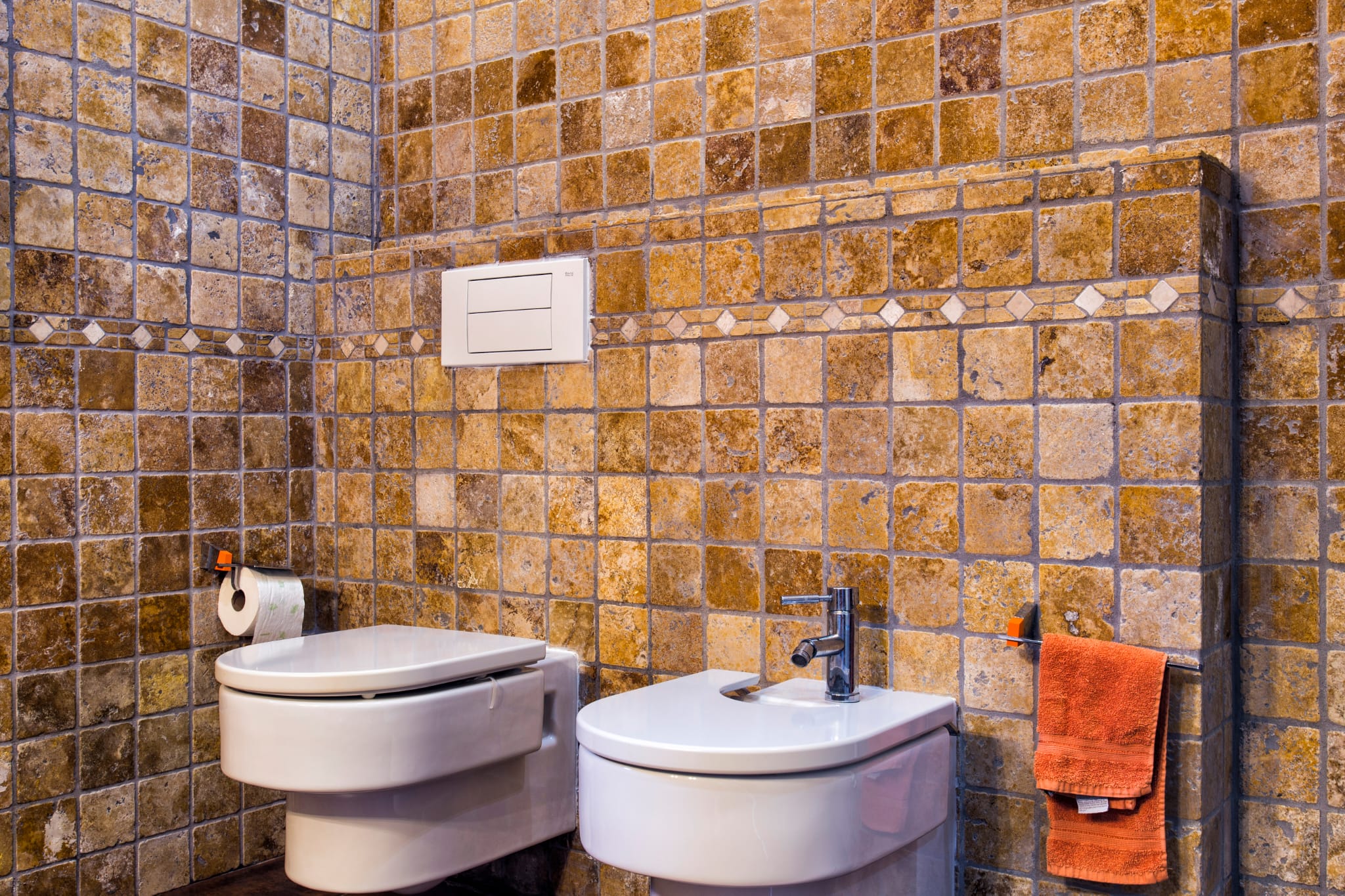 Salle de bain avec carrelage rustique aux tons terreux, WC et bidet suspendus en céramique blanche et frise décorative.