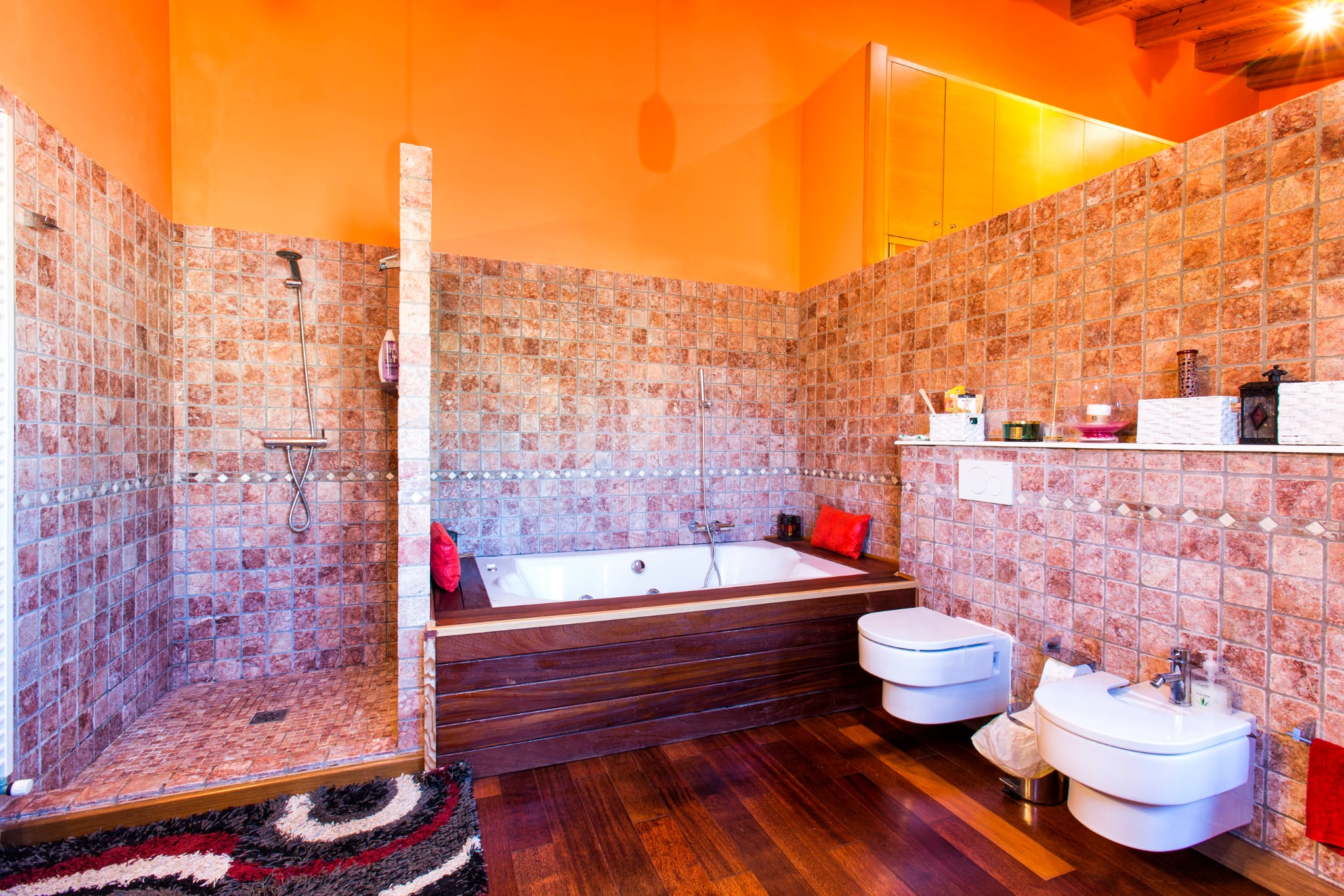 Salle de bain avec douche à l'italienne en pierre, baignoire habillée de bois sombre et sanitaires suspendus. Murs orange et parquet en bois chaleureux sous plafond boisé.
