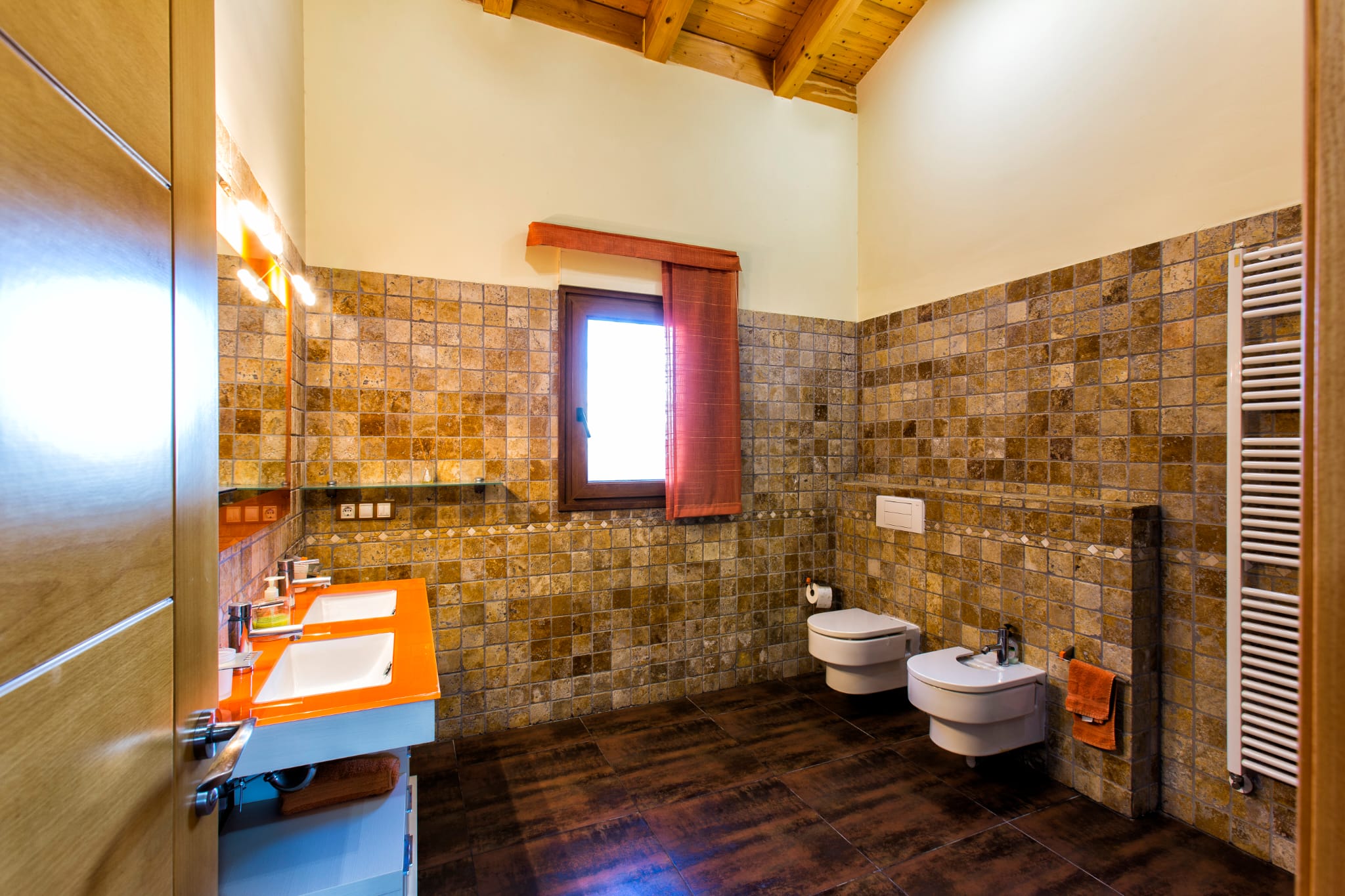 Salle de bain moderne avec double vasque orange, murs en pierre naturelle et plafond voûté en bois avec poutres apparentes.