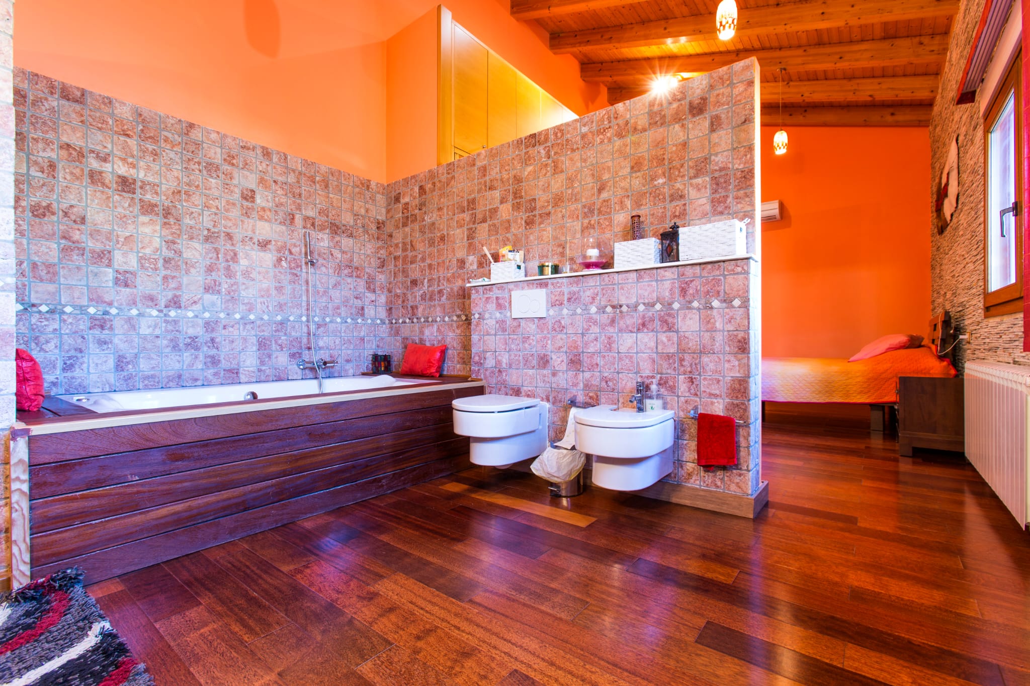 Salle de bain ouverte avec baignoire habillée de bois, murs en mosaïque de pierre et sanitaires suspendus, intégrée à une chambre sous un plafond de poutres apparentes.