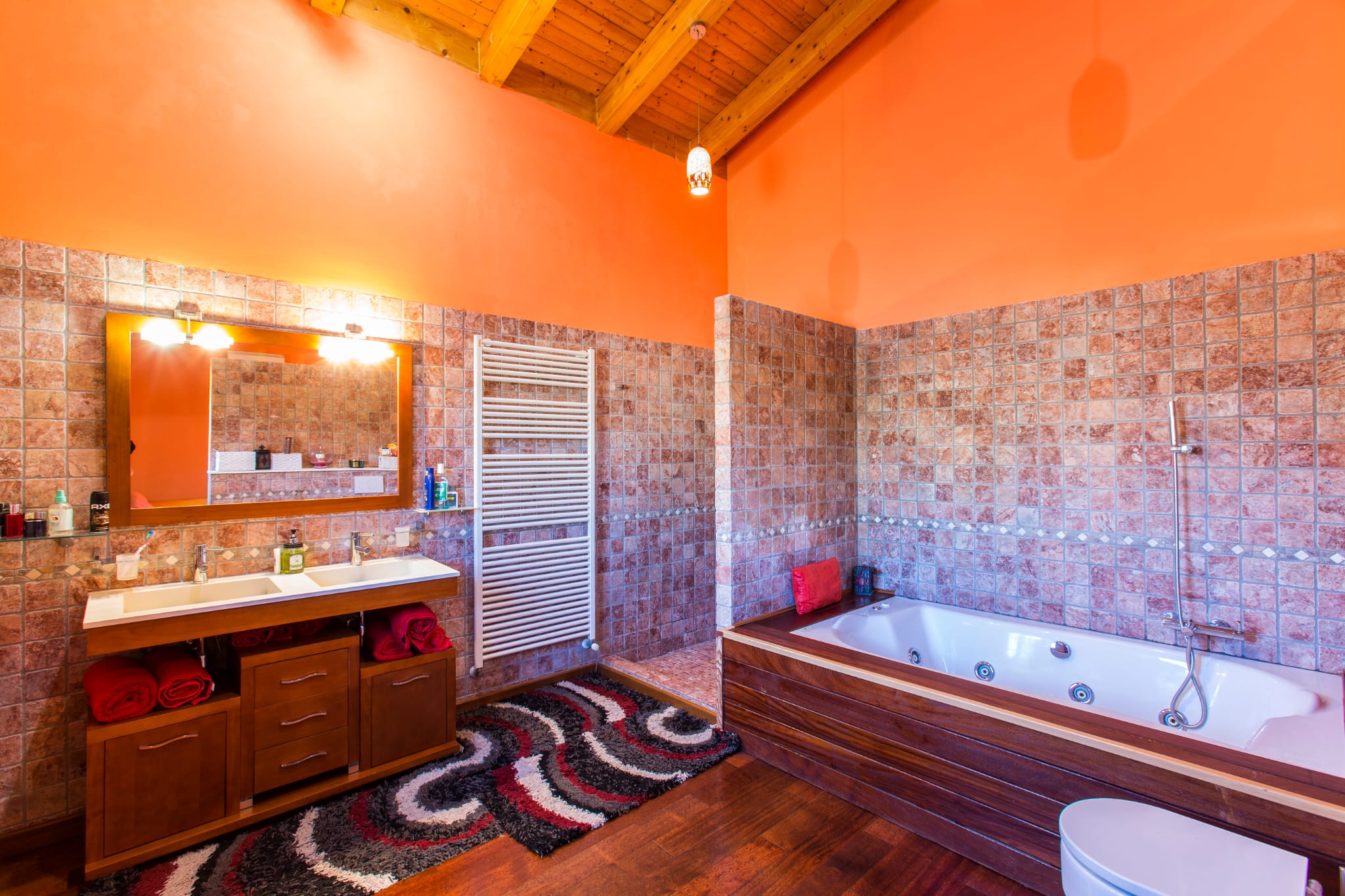 Salle de bain rustique avec poutres apparentes, double vasque sur meuble en bois foncé, baignoire balnéo et murs carrelés dans les tons terre cuite et orange.