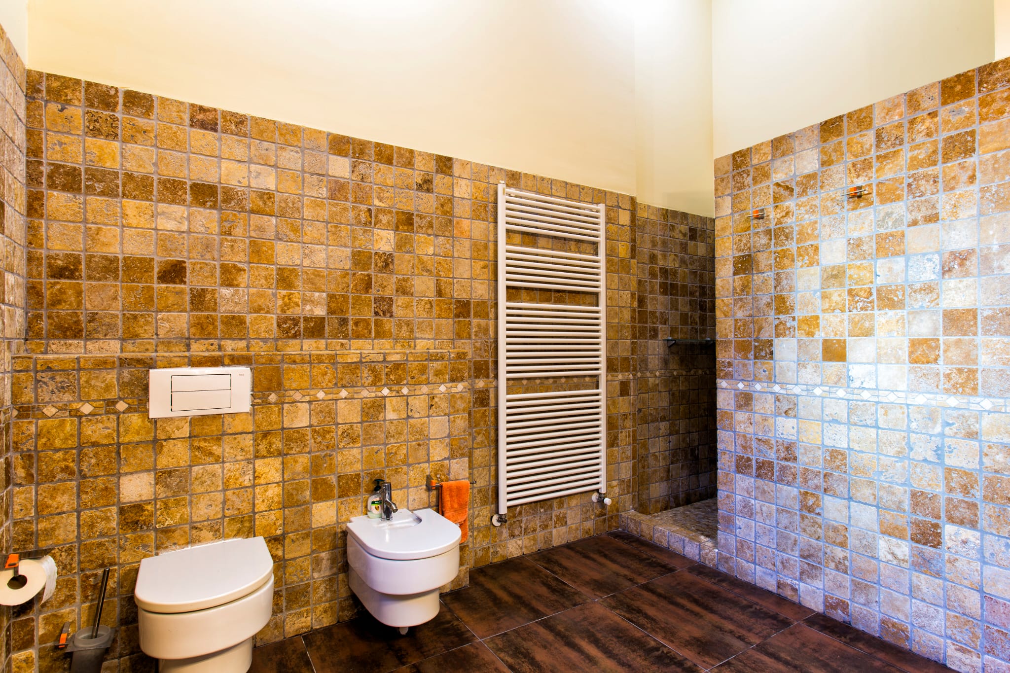 Salle de bains avec murs en mosaïque de pierre naturelle, toilettes et bidet suspendus, sèche-serviettes blanc et sol en grès cérame aspect bois.