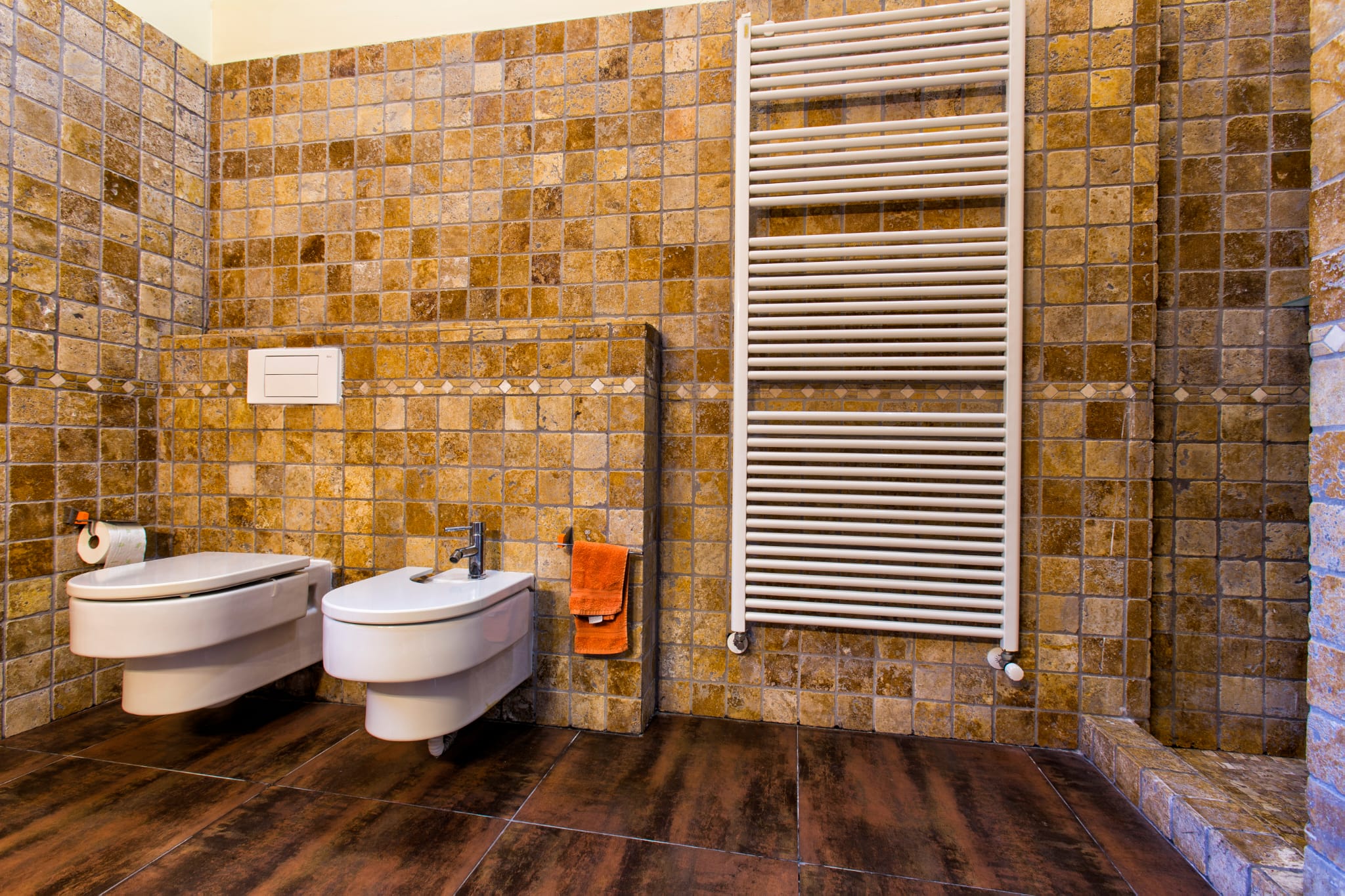 Salle de bains avec toilettes et bidet suspendus, murs en mosaïque de pierre aux tons ocres et grand radiateur sèche-serviettes blanc.