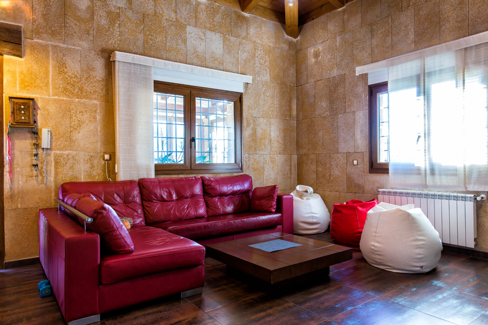 Salon avec canapé d'angle en cuir rouge, murs en pierre naturelle et poutres apparentes. Comprend une table basse en bois et des poufs d'appoint.