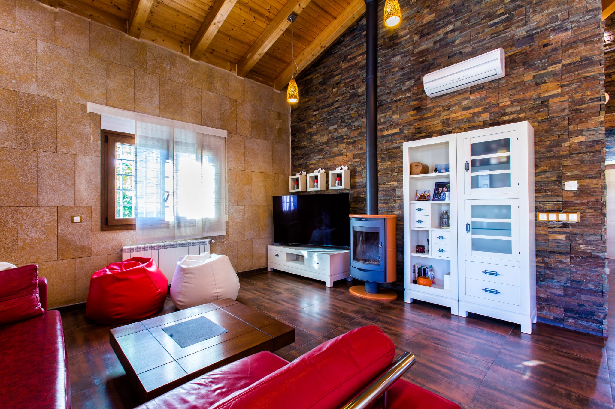 Salon rustique avec murs en pierre naturelle, plafond en poutres de bois, poêle à bois moderne et canapés en cuir rouge dans un espace chaleureux.