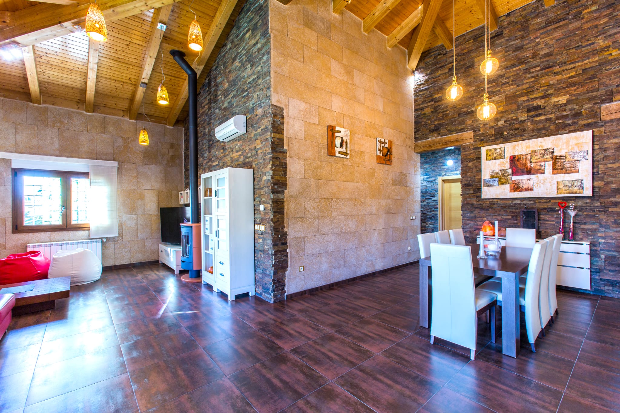 Salon-salle à manger avec plafonds voûtés en bois, murs en pierre naturelle et poêle à bois. L'espace dispose de luminaires suspendus et d'un sol en grès cérame foncé.
