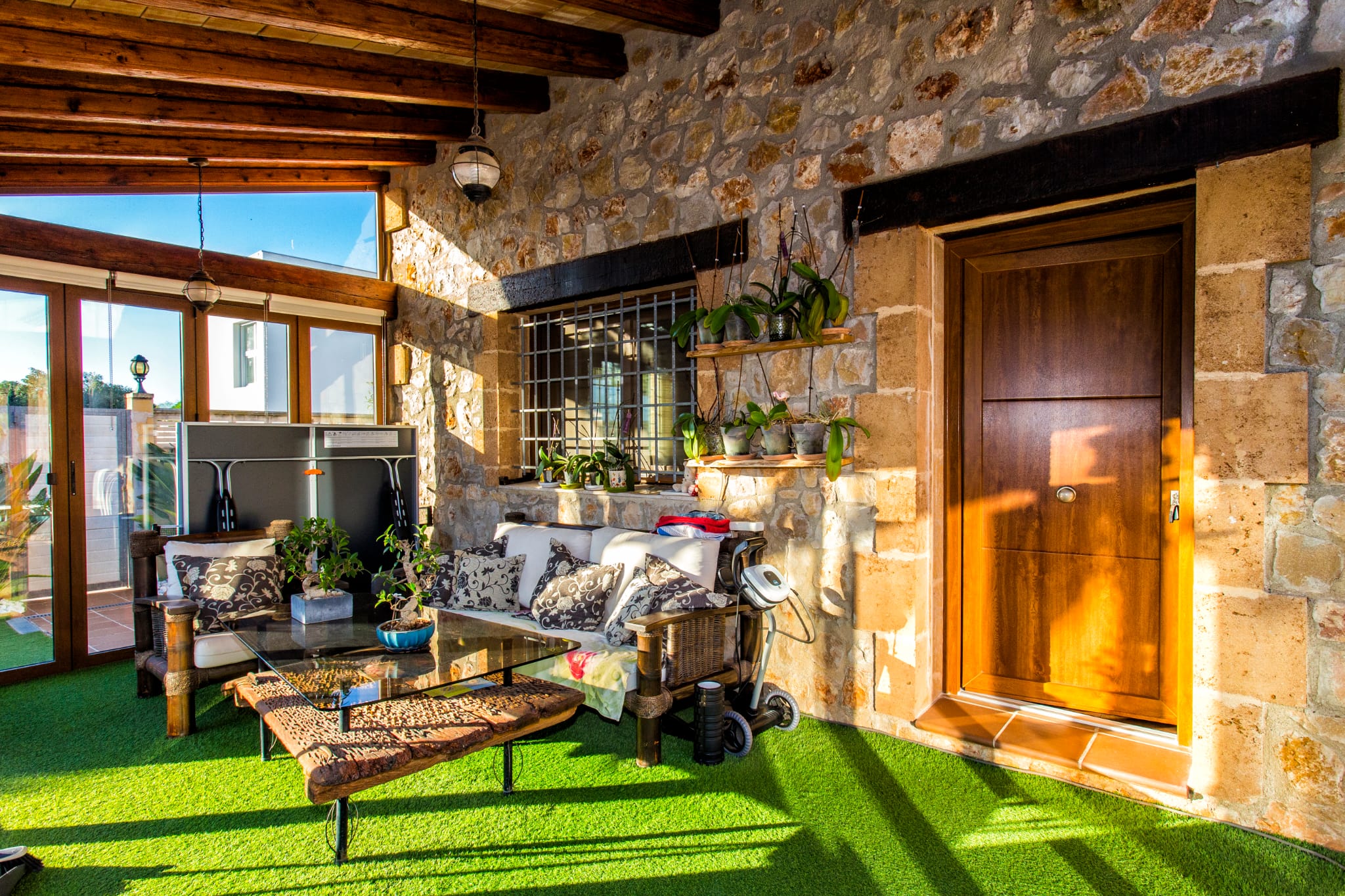Terrasse couverte avec murs en pierre apparente, poutres en bois et gazon synthétique. Espace salon avec table en bois flotté et étagères d'orchidées.