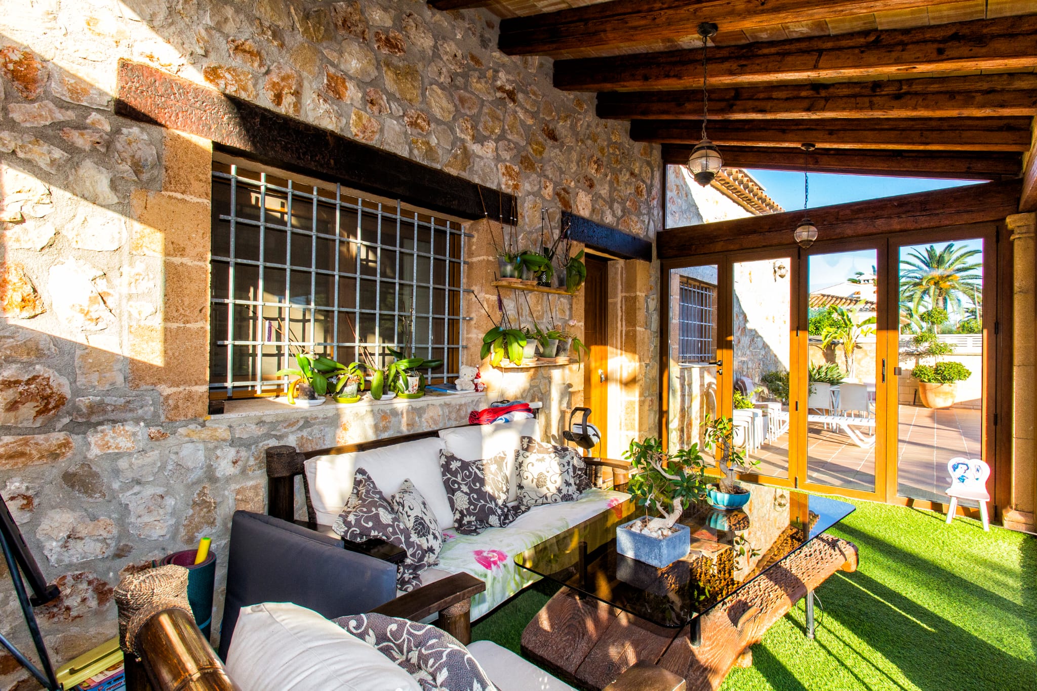 Terrasse couverte avec murs en pierre apparente, poutres en bois sombre et coin salon avec table en verre sur gazon synthétique, ouvrant sur un patio ensoleillé.