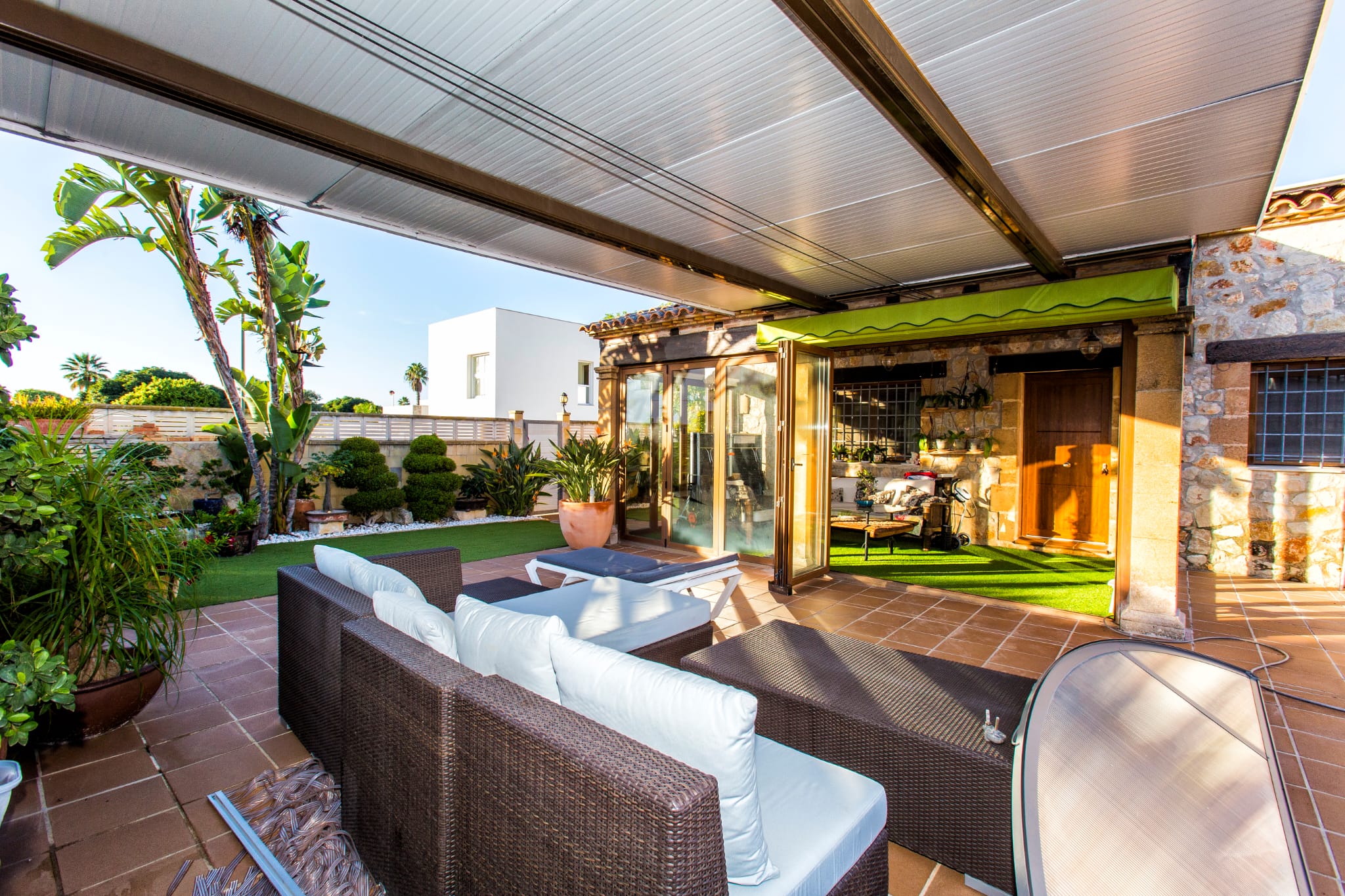 Terrasse couverte équipée d'un mobilier en résine tressée, sol en grès et jardin avec gazon synthétique bordé de plantes tropicales et murs en pierre.