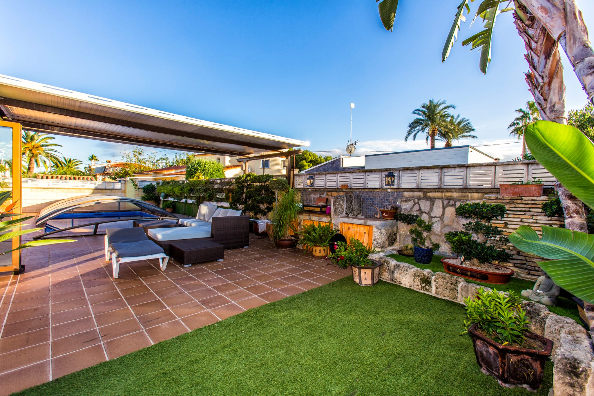 Terrasse extérieure avec carrelage en terre cuite, gazon synthétique et salon sous pergola, à côté d'une piscine avec abri télescopique et murets en pierre.