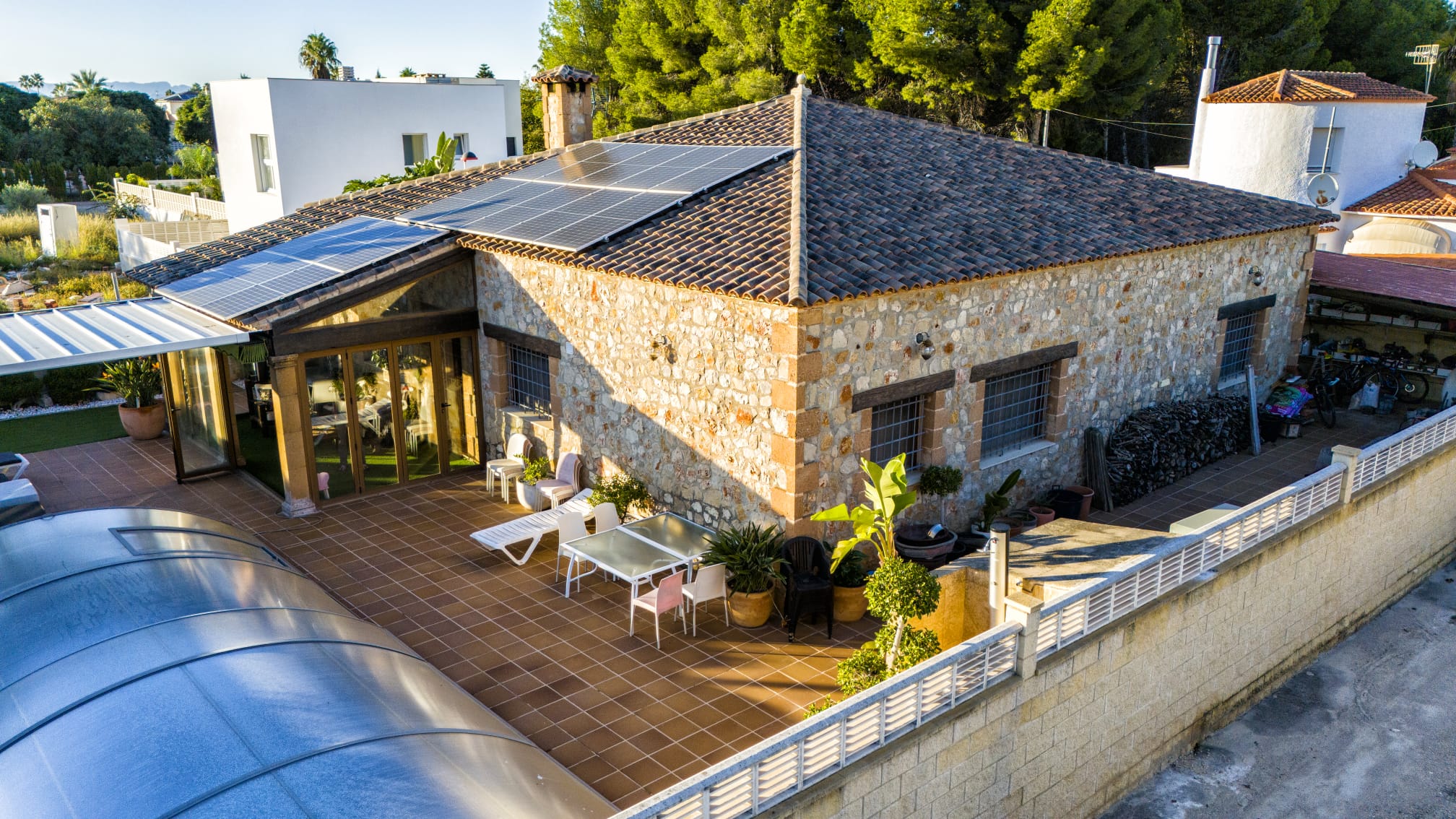 Vue extérieure d'une villa en pierre avec panneaux solaires, vaste terrasse carrelée et piscine dotée d'un abri télescopique.