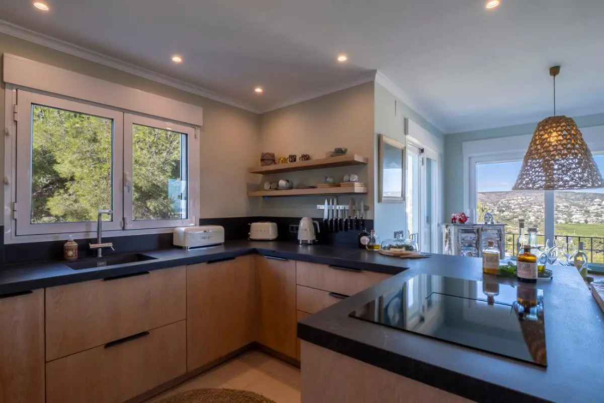 Cuisine moderne avec des meubles en bois clair, des plans de travail sombres et une plaque à induction. Les grandes fenêtres offrent une vue sur les pins et la vallée.