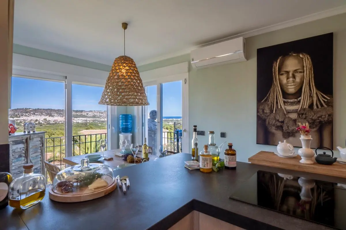 Cuisine moderne avec plan de travail sombre, plaque à induction et coin repas éclairé par une suspension en osier, offrant une vue panoramique sur la côte.