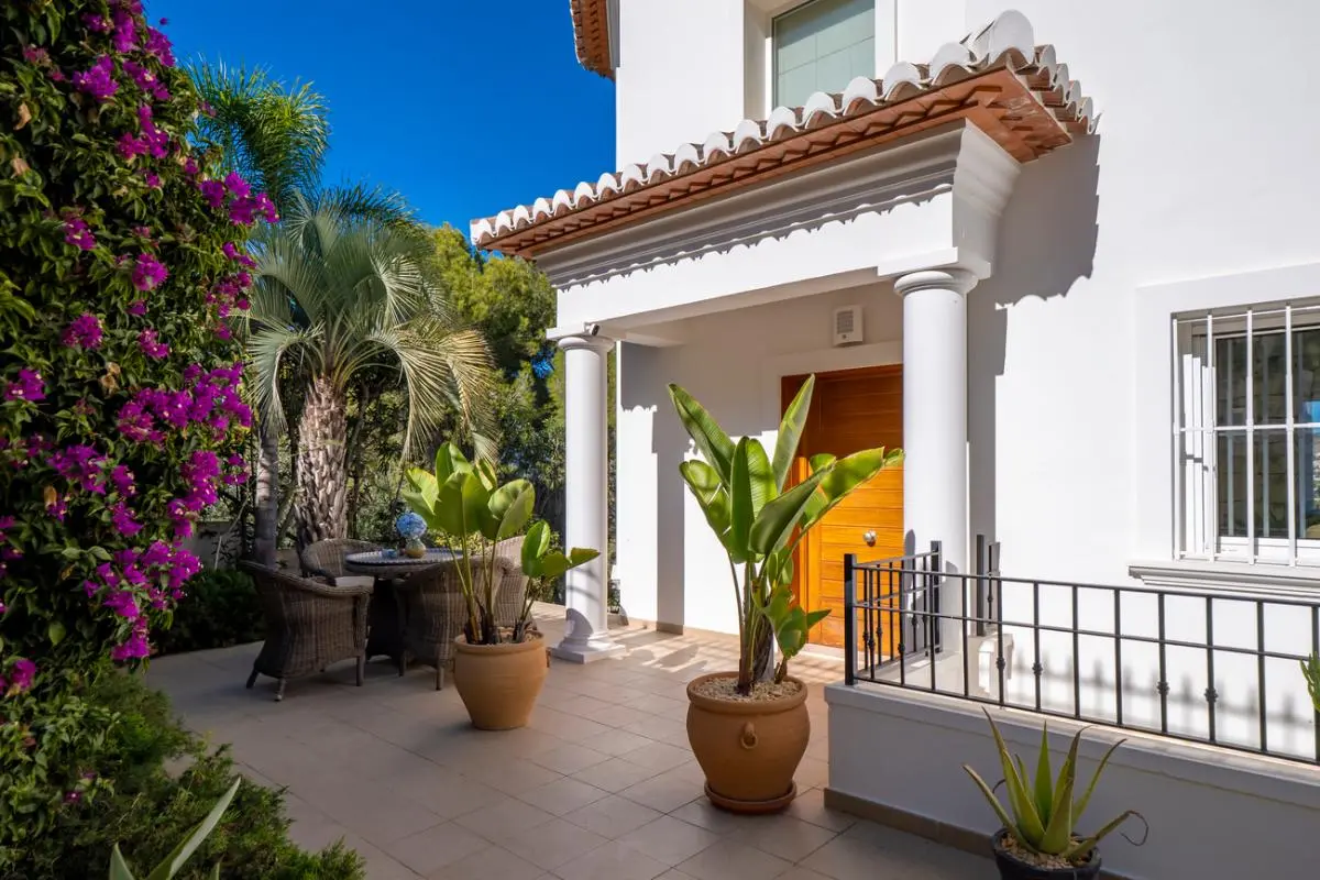 Entrée principale avec porche à colonnes blanches, porte en bois et terrasse carrelée entourée de bougainvilliers et de plantes tropicales.
