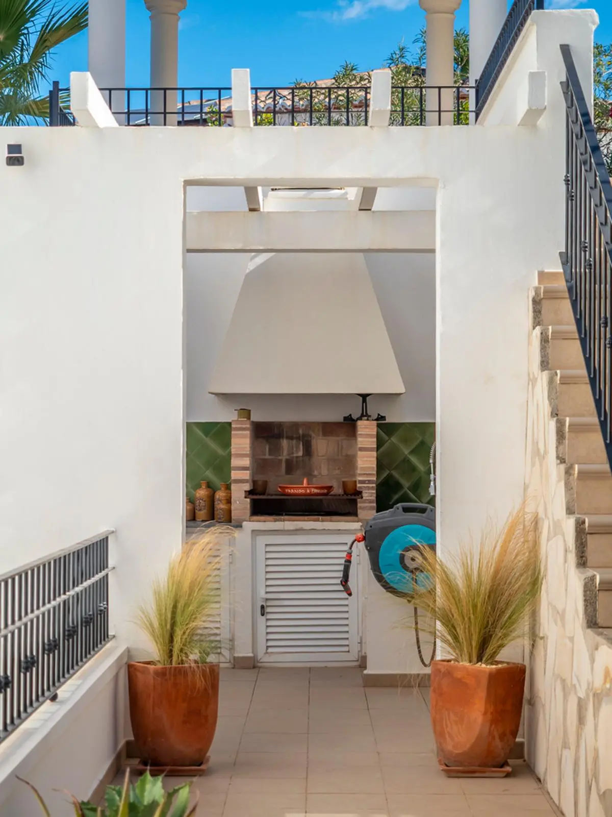 Espace barbecue extérieur avec grill en briques, hotte blanche et crédence en carreaux verts, encadré par des jardinières en terre cuite et des murs blancs.