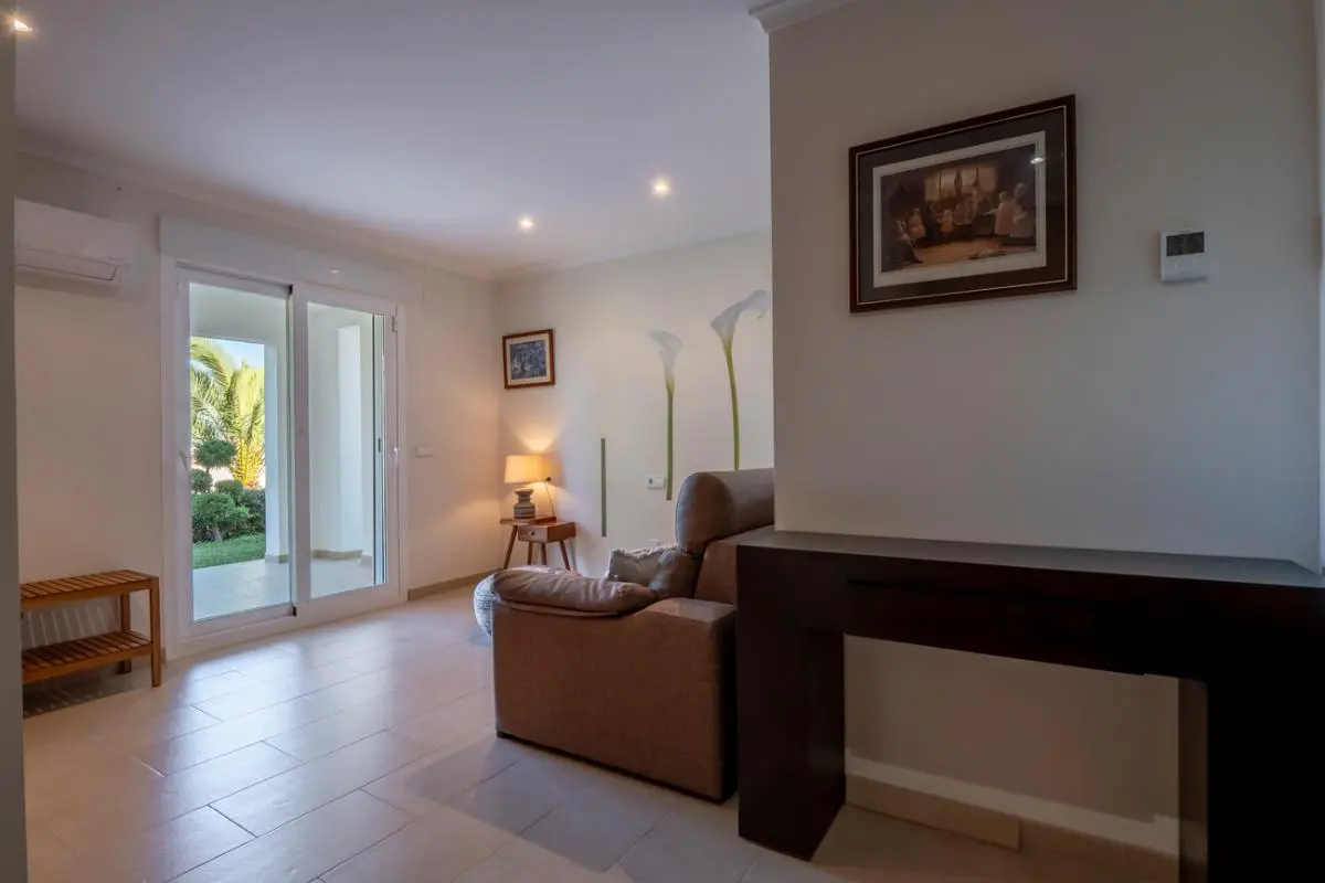 Espace de vie avec console en bois sombre, fauteuil en cuir marron et carrelage clair. Une baie vitrée coulissante ouvre sur le jardin et les palmiers.