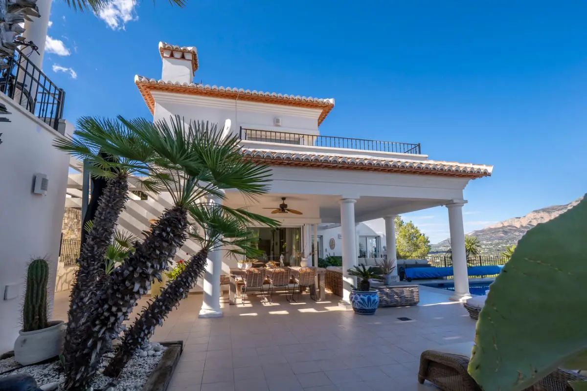 Extérieur d'une villa blanche avec terrasse couverte à colonnes, espace repas extérieur et jardin méditerranéen offrant une vue sur les montagnes.