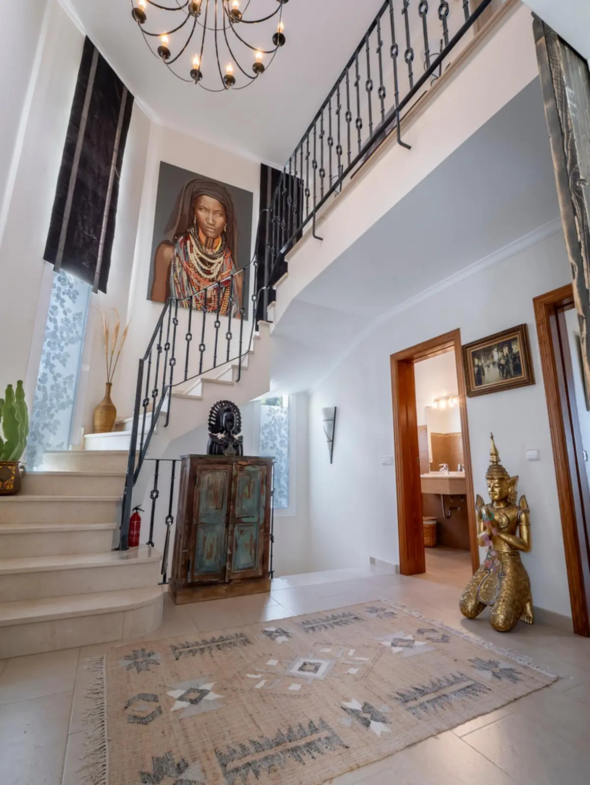 Hall d'entrée avec escalier en marbre, rampe en fer forgé, grand portrait mural et décoration éclectique incluant une statue dorée et un tapis à motifs.