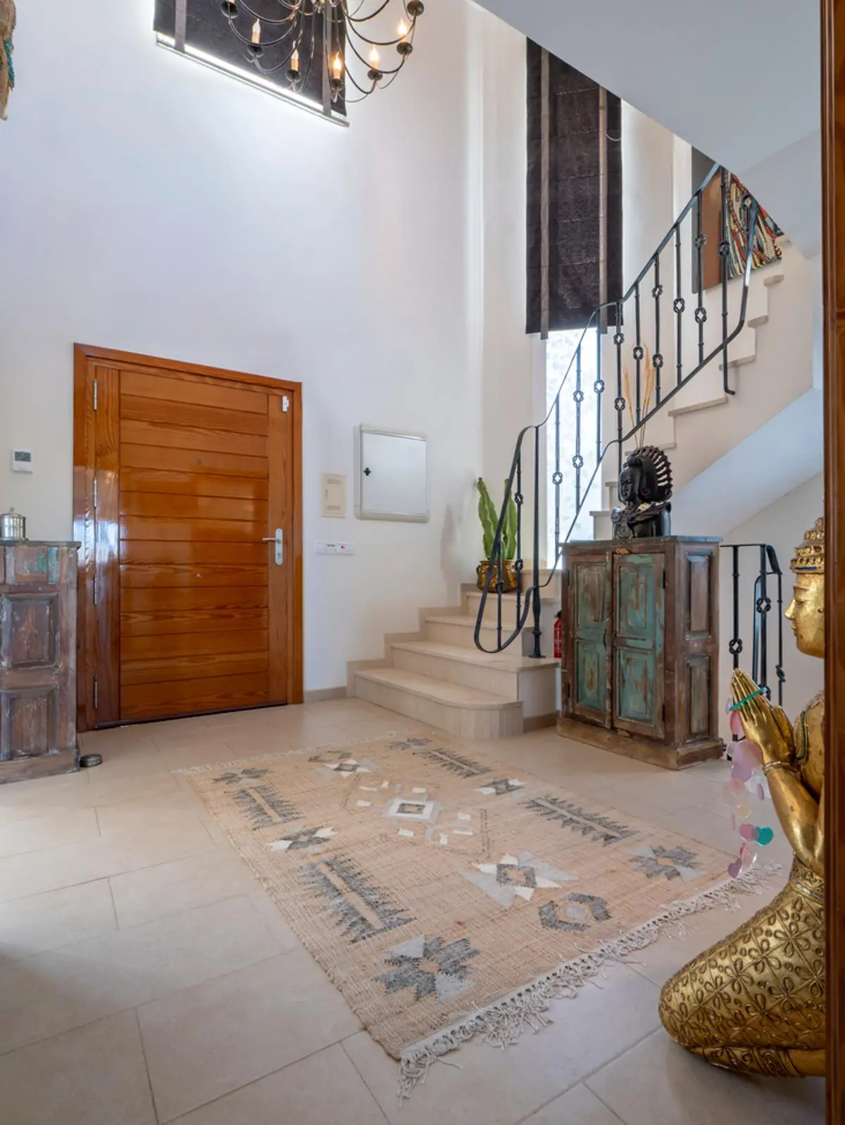 Hall d'entrée avec porte en bois massif, escalier en pierre à rampe en fer forgé et haut plafond. Décoration rustique, lustre classique et sol en carrelage clair.