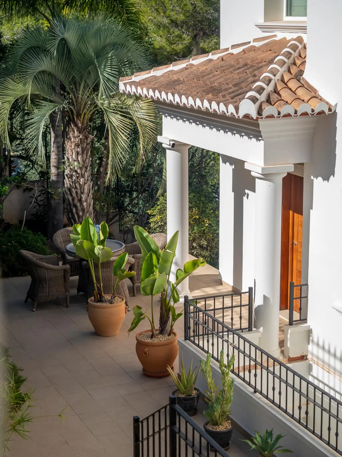 Porche d'entrée aux colonnes blanches et toit en terre cuite. Le patio dallé dispose de plantes tropicales en pots, de balustrades en fer forgé et d'une porte en bois.