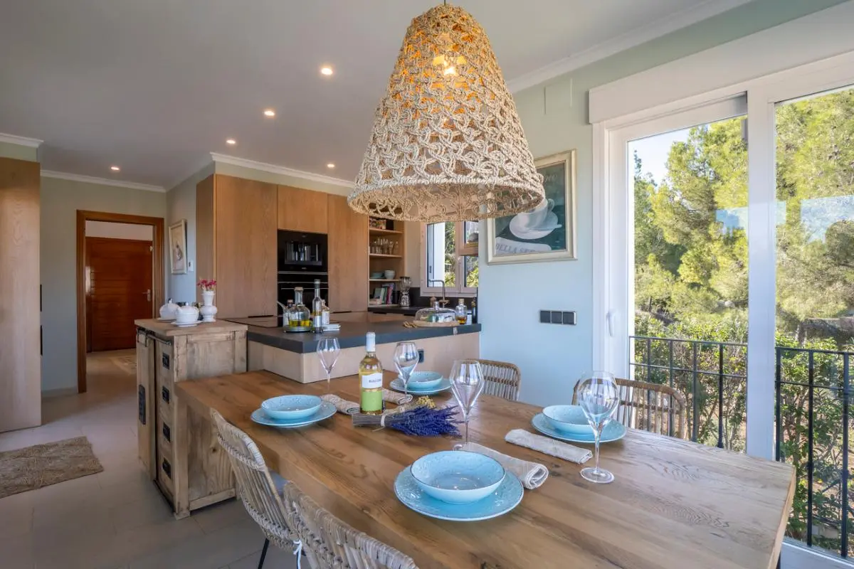 Salle à manger avec table en bois, suspension en osier et cuisine intégrée en chêne. Les baies vitrées s'ouvrent sur un environnement verdoyant et paisible.