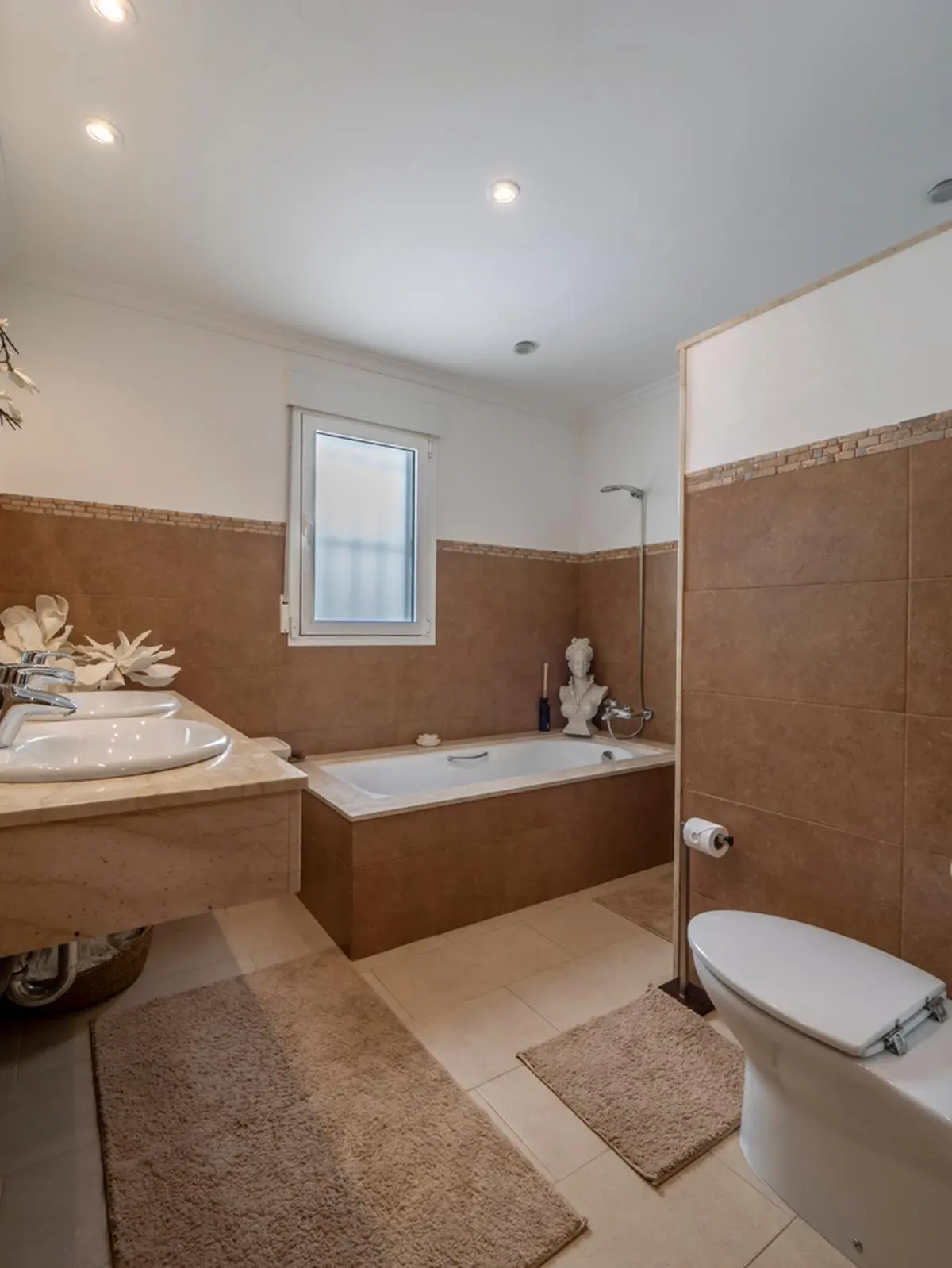 Salle de bain avec double vasque sur plan en marbre, baignoire encastrée en grès brun et éclairage par spots intégrés au plafond.