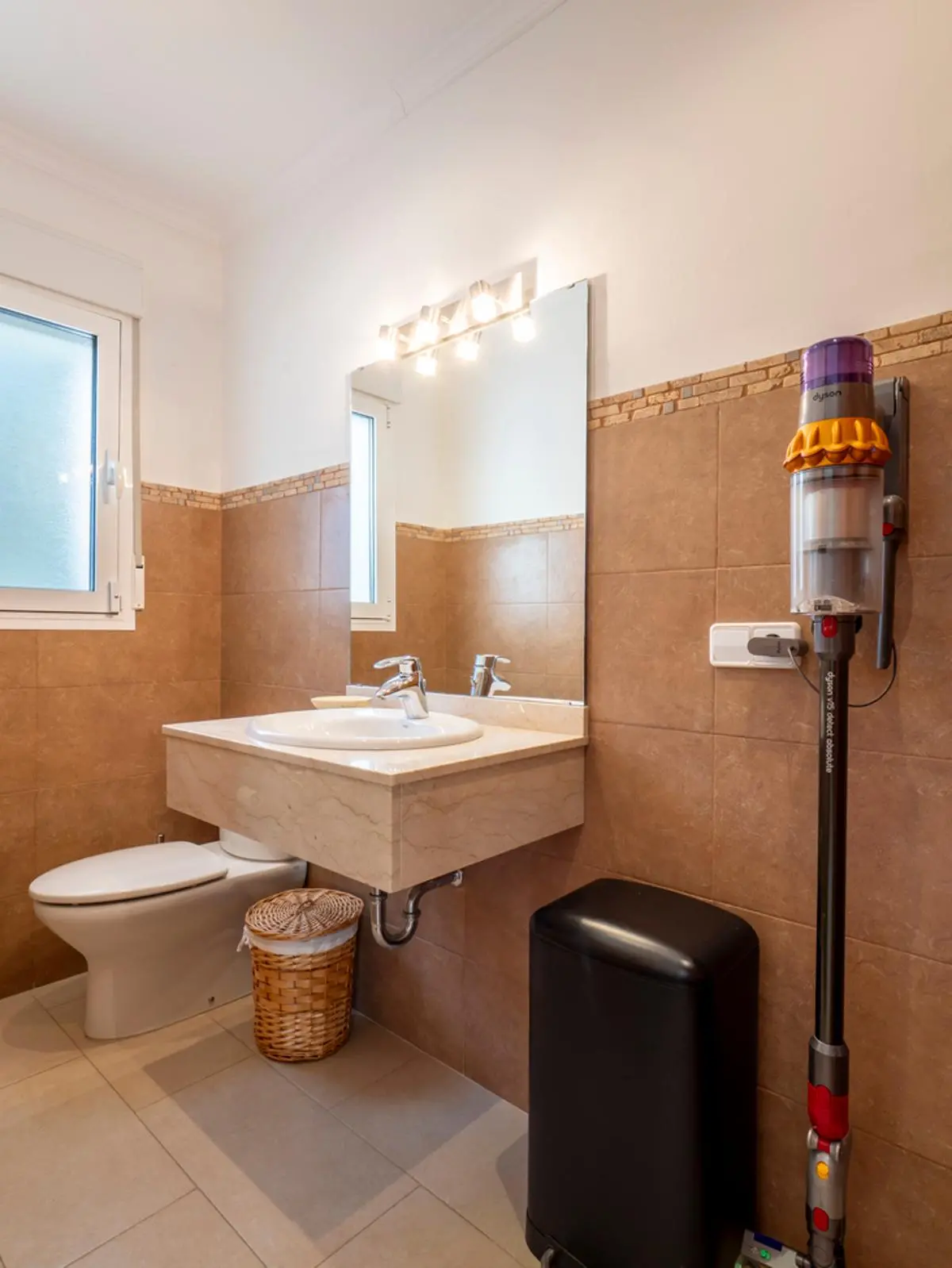 Salle de bain avec meuble vasque suspendu en marbre, carrelage beige et miroir éclairé. La pièce comprend une fenêtre dépolie et un aspirateur Dyson mural.