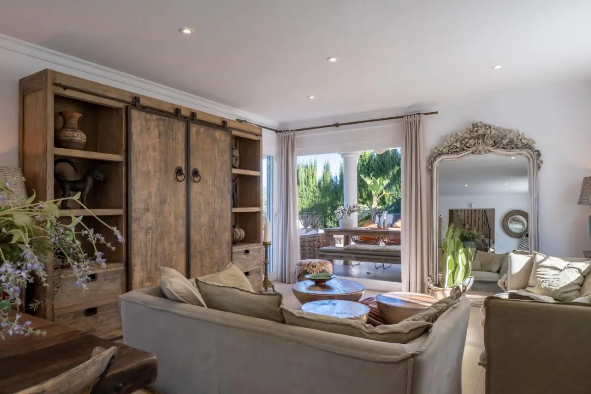 Salon avec un grand meuble en bois rustique, des canapés neutres et un miroir ornementé, s'ouvrant sur une terrasse avec espace de repas extérieur.