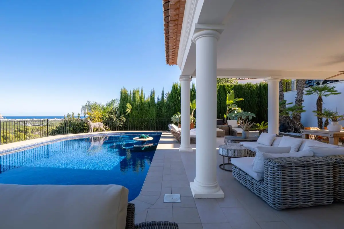 Terrasse couverte avec colonnes blanches et mobilier en rotin, bordant une piscine en mosaïque avec vue panoramique sur la côte.