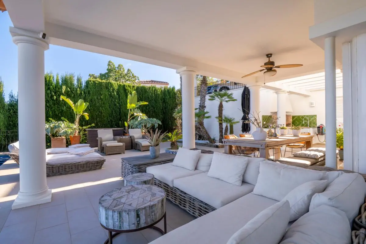 Terrasse couverte avec colonnes blanches, grand canapé d'angle en osier et espace repas extérieur avec ventilateur, bordée de plantes tropicales.