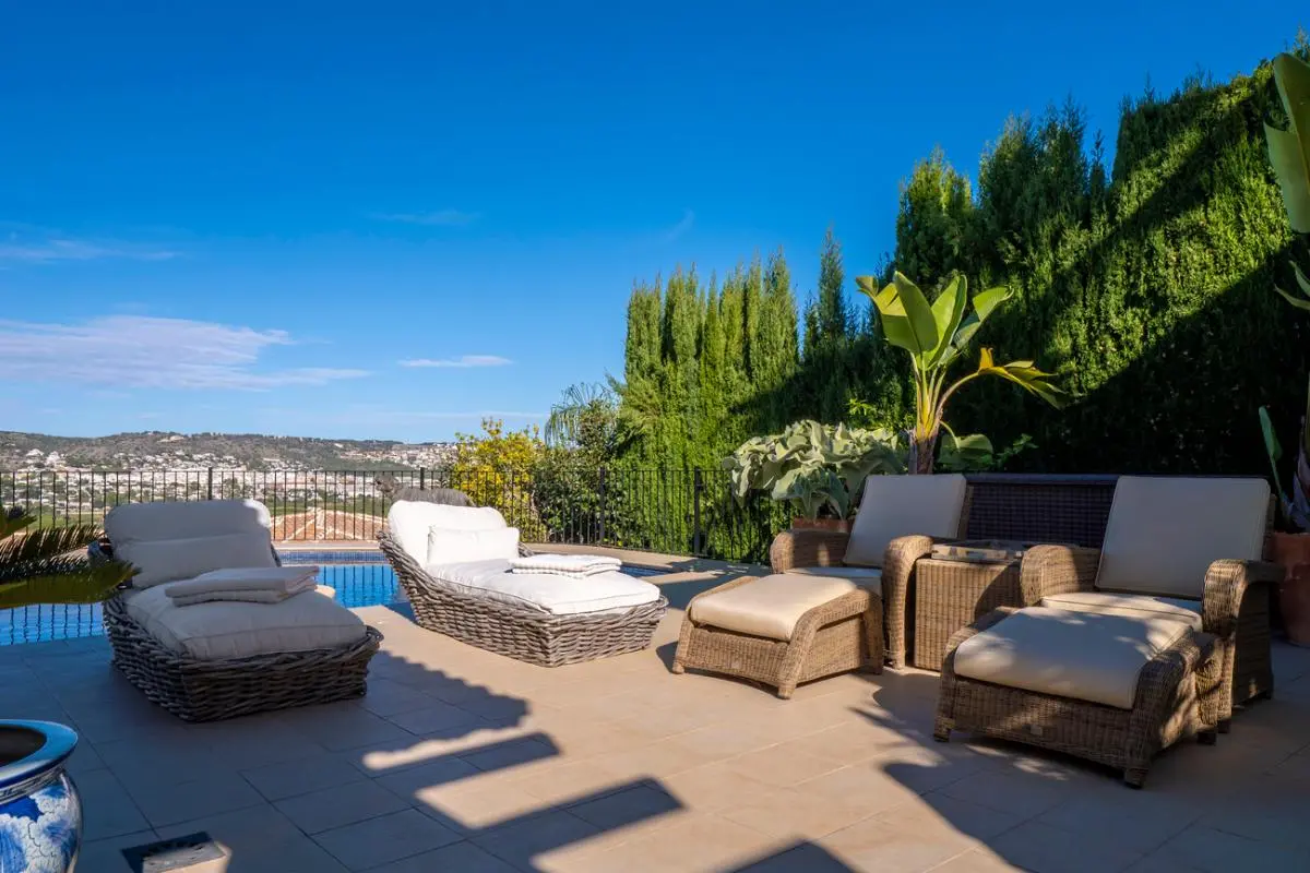 Terrasse équipée de bains de soleil en rotin et coussins blancs, offrant une vue sur les collines et bordée d'une haie de cyprès.