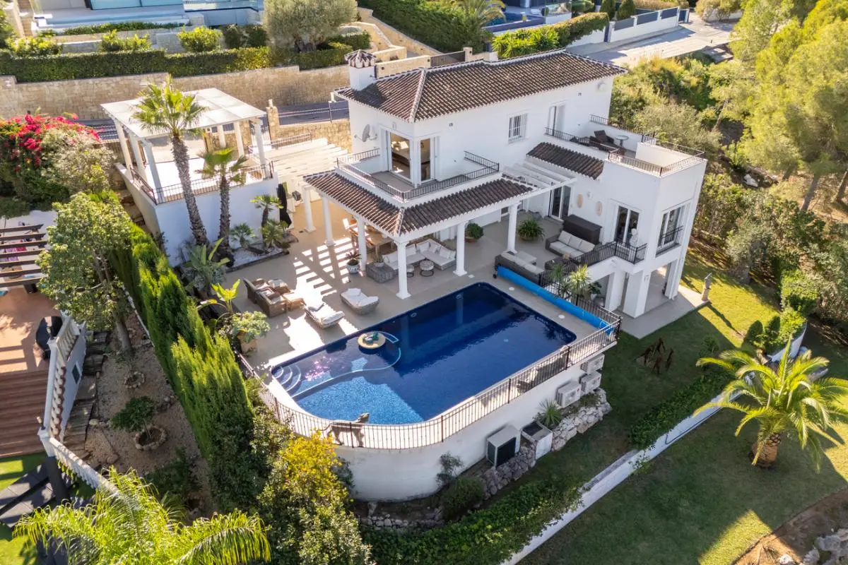 Vue aérienne d'une villa méditerranéenne blanche avec piscine, plusieurs terrasses couvertes à colonnes et jardins paysagers avec palmiers.